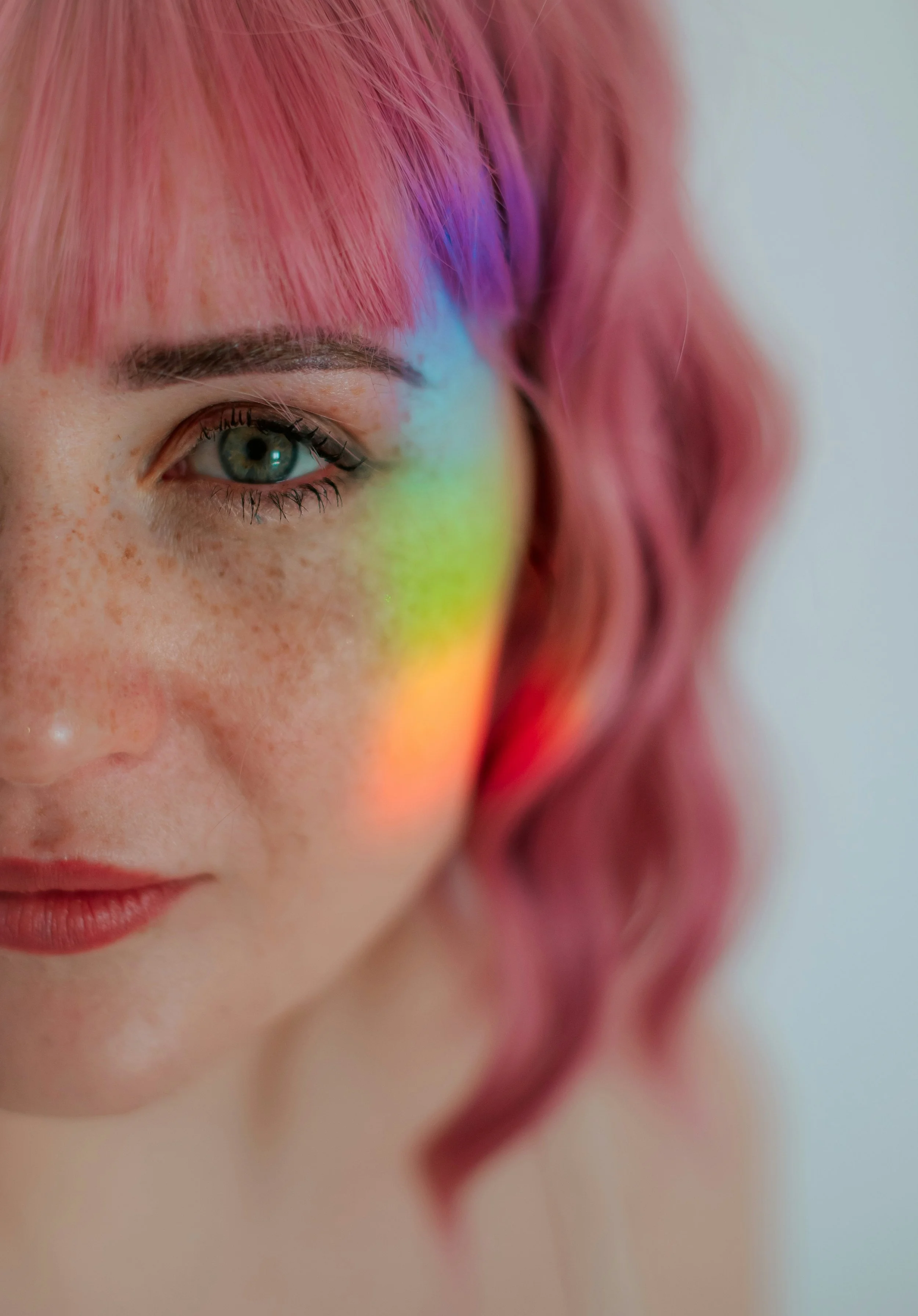 Close-up of a person's face showing one eye, pink hair, freckles, light makeup, and a rainbow-colored reflection on the skin.
