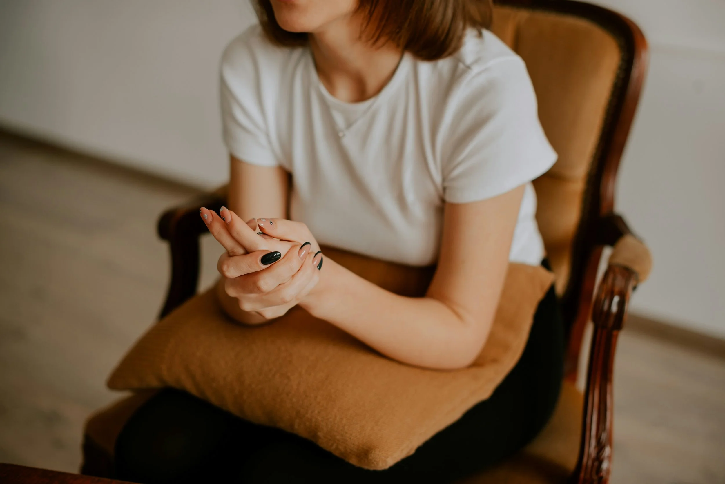 Woman with red hair wearing a white shirt sitting on a wooden chair with armrests, hands clasped together, in a room with beige walls.