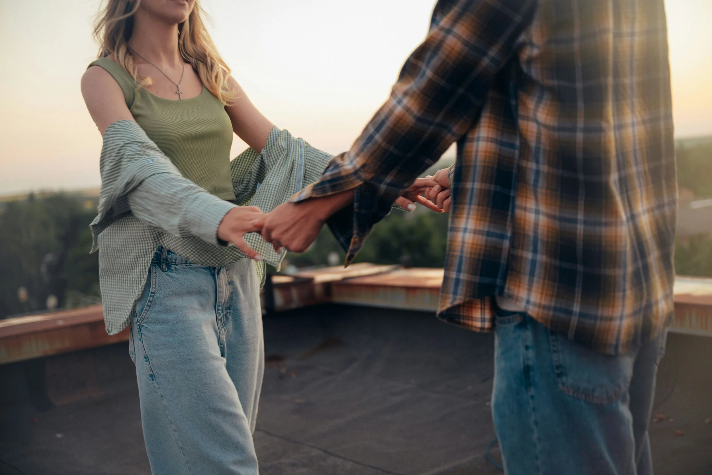 A couple holding hands, standing outdoors during sunset. The woman is wearing a green tank top, high-waisted jeans, and a necklace with a cross pendant. The man is wearing a plaid shirt. The background shows a blurred landscape and a setting sun.