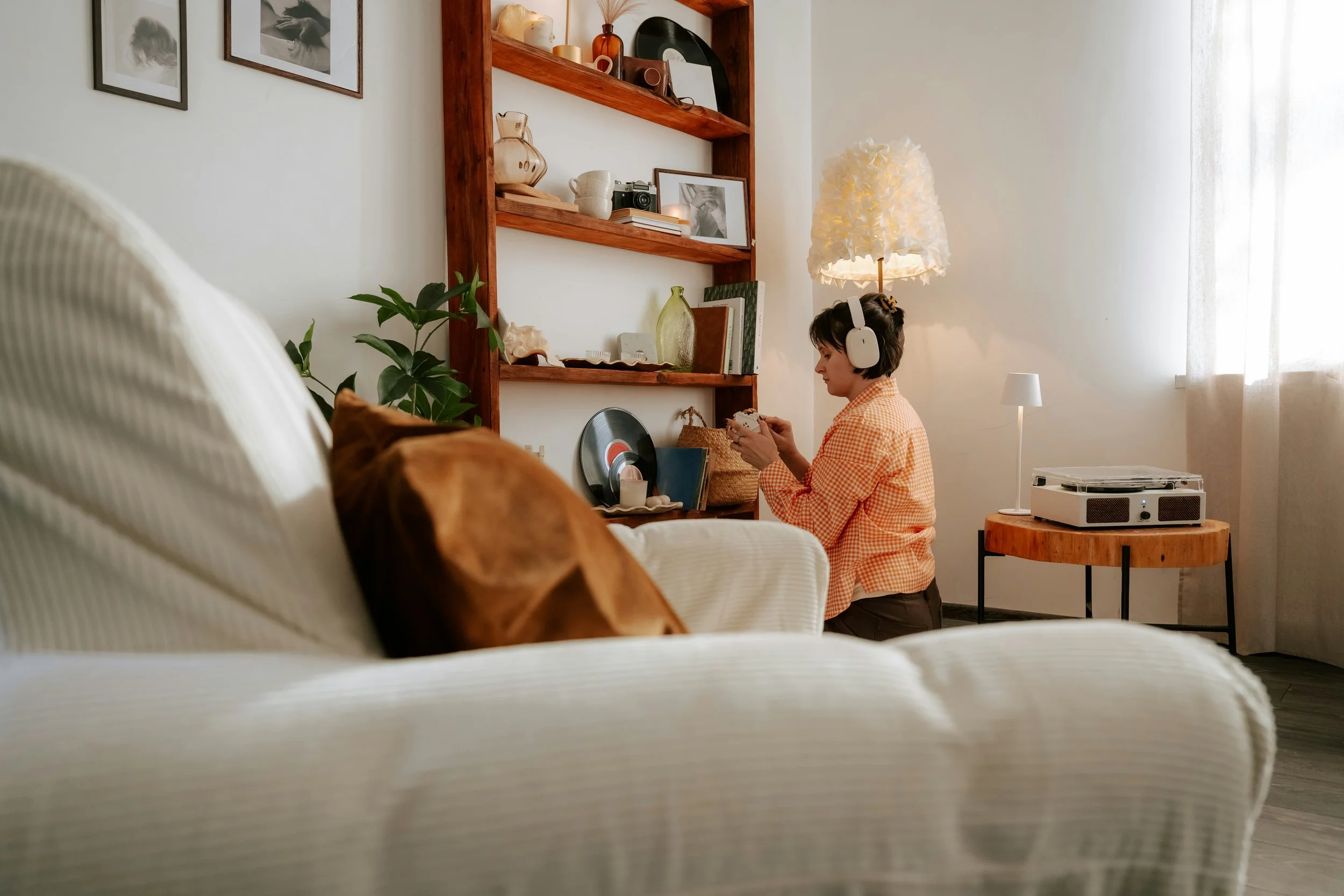 A woman with short dark hair, wearing a red and white checkered shirt, sits on the floor with headphones on, holding a vinyl record. The room has white walls, a wooden shelf with decorations and framed photos, a lamp with a floral shade, and a small table with a vintage record player. A dog is partially visible in the foreground, and a plant is on the left.