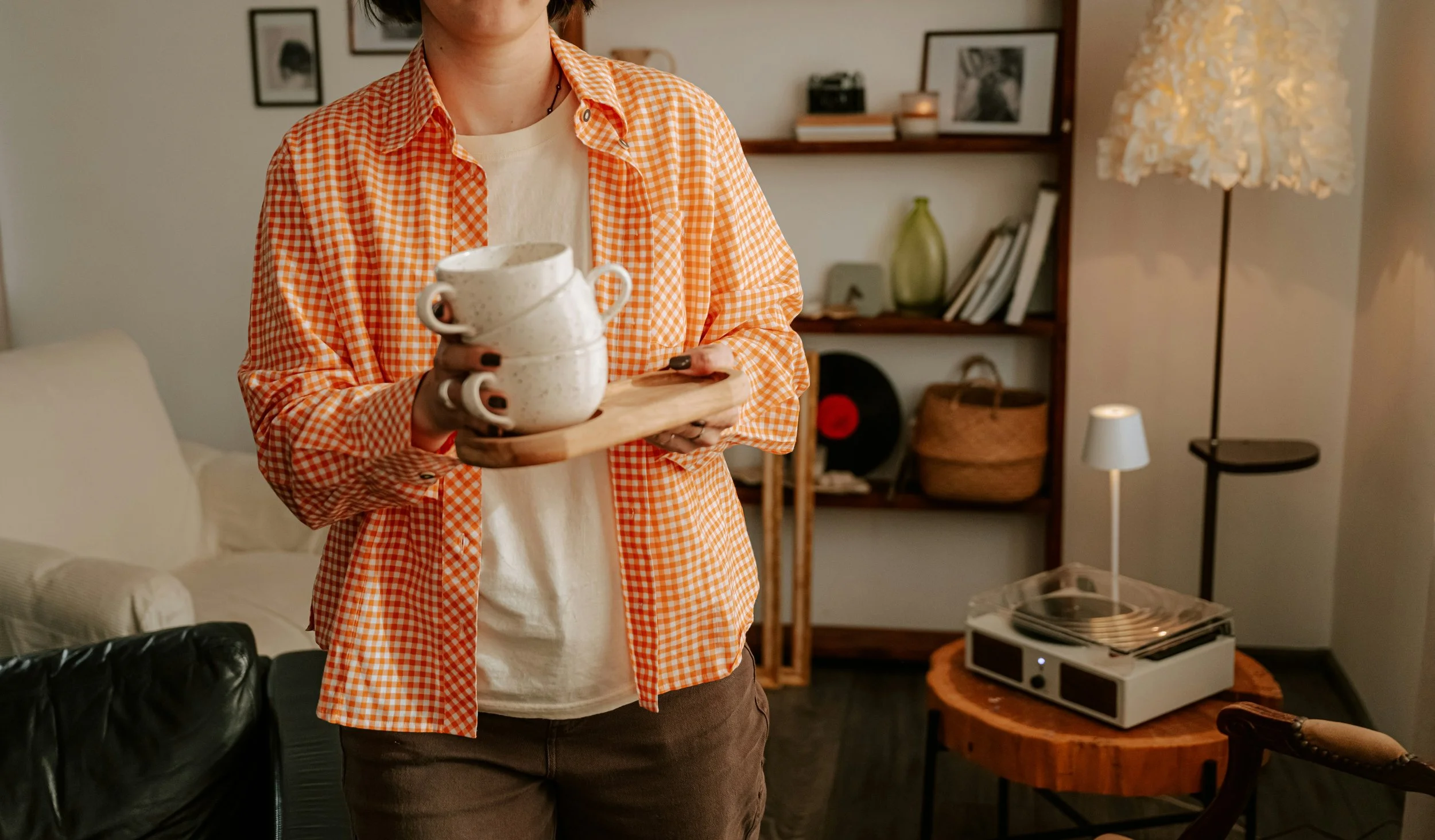 A woman is holding a wooden tray with three stacked teacups in a cozy living room with bookshelf, artwork, and a floor lamp.