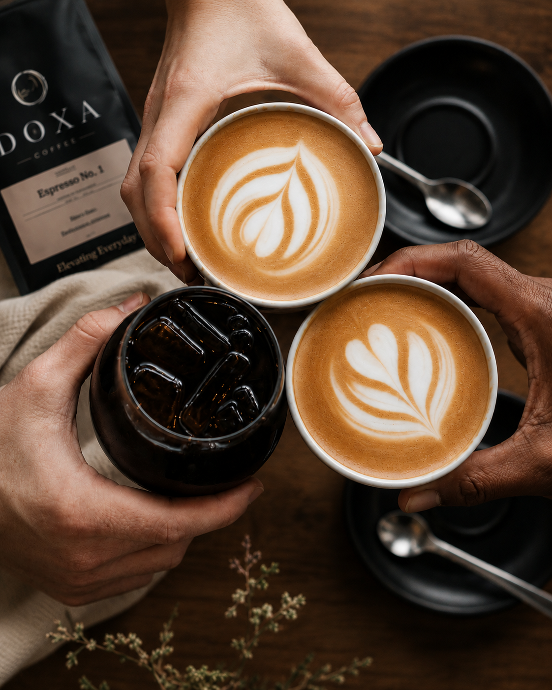 Three people toasting with coffee drinks, two cappuccinos with latte art and a glass of iced coffee on a wooden table.