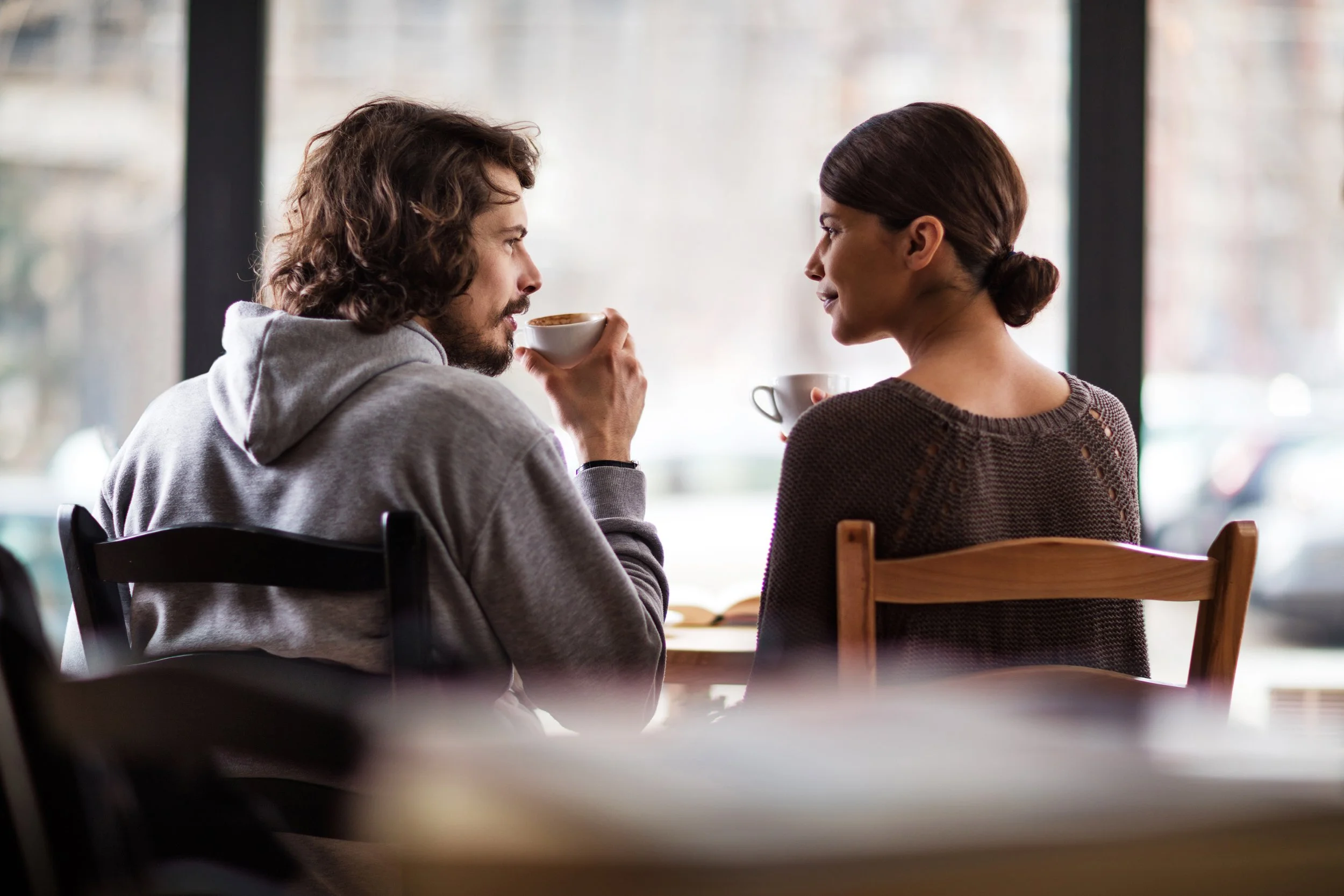 Two people sitting at a table in a cafe, drinking coffee and facing each other, with large windows in the background.