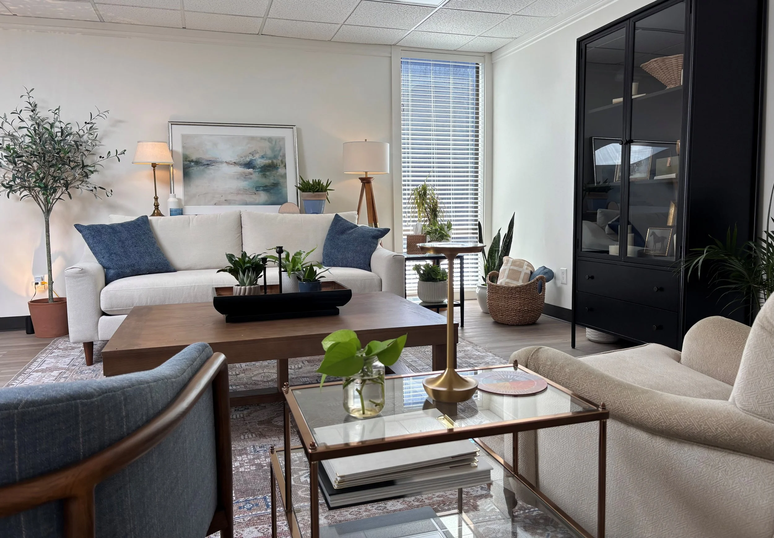 A cozy living room with a white sofa and navy blue pillows, a wooden coffee table with a glass top and decorative items, a black cabinet, potted plants, a beige armchair, and large windows with white blinds letting in natural light.