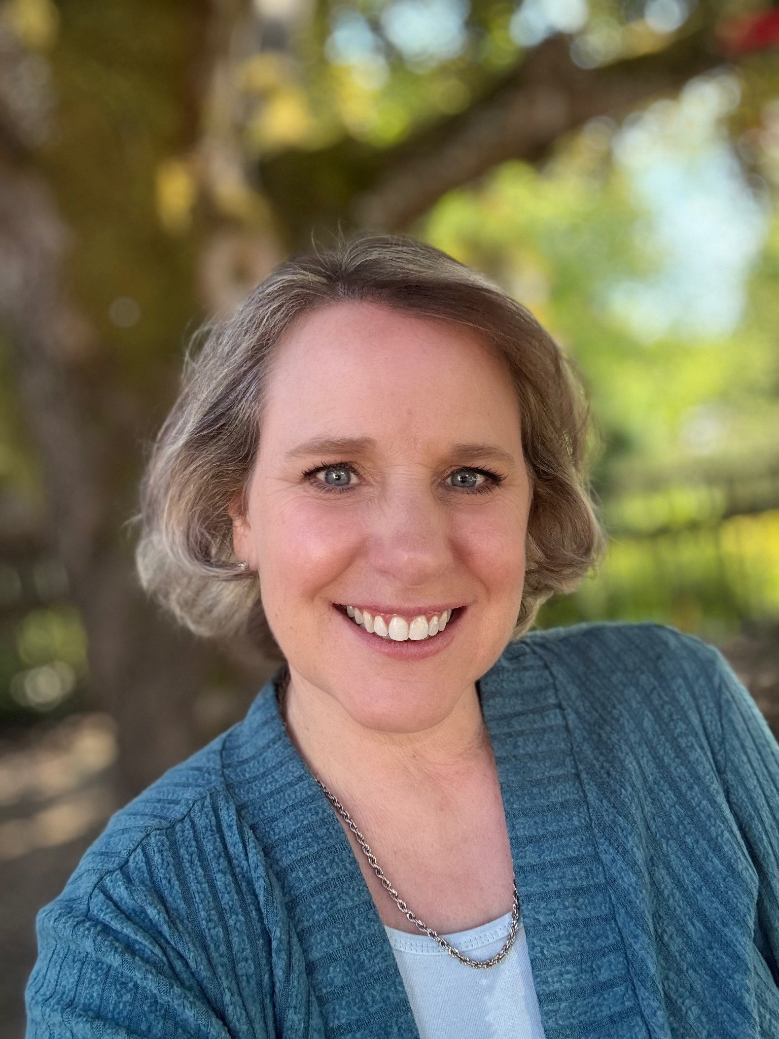 A woman with short gray hair and blue eyes smiling outdoors, wearing a blue jacket, white top, and a silver necklace, with a background of blurred trees and sunlight.