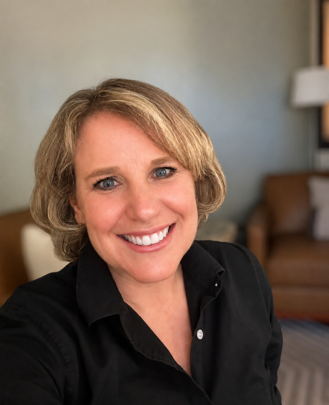 A smiling woman with short blond hair, wearing a black shirt, sitting in a cozy room with a brown sofa and lamp in the background.