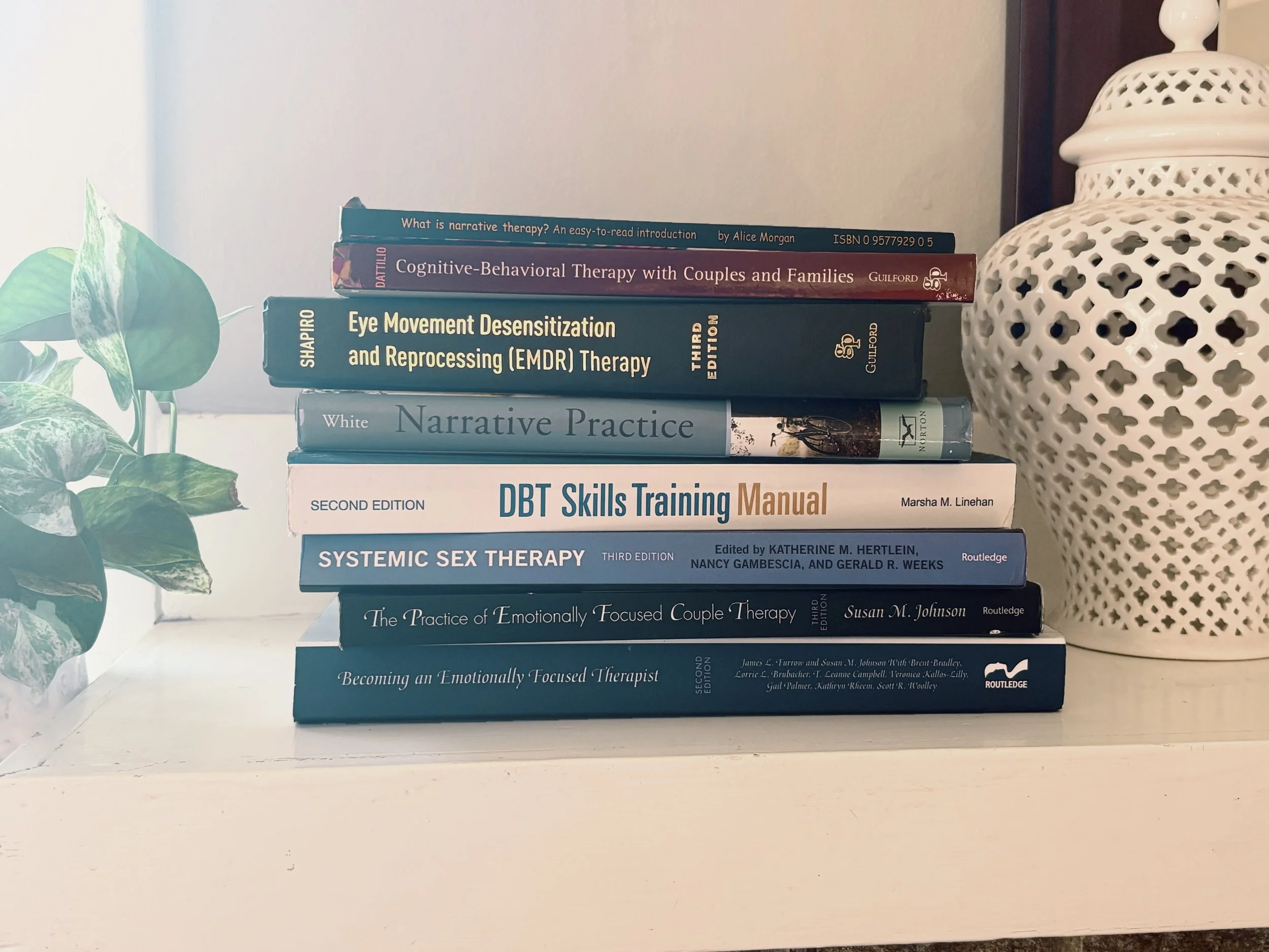 Stack of psychotherapeutic books on therapy techniques and methods on a white shelf, with a white lattice-patterned vase to the right and a green leafy plant to the left.