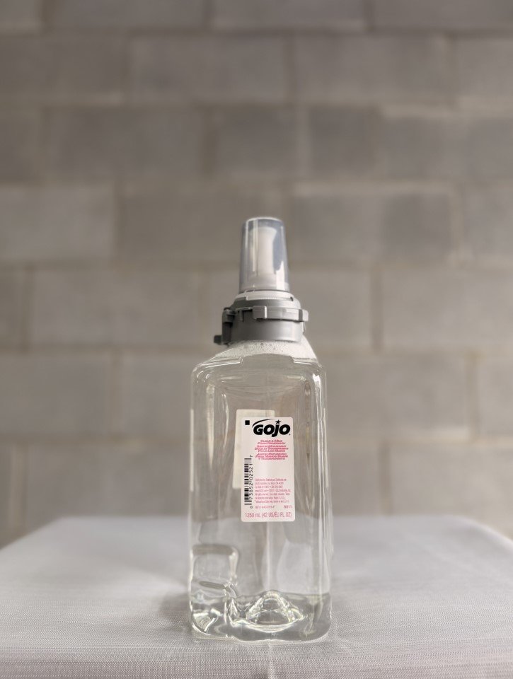 A clear plastic spray bottle with a white label and black and pink text, standing on a white surface with a brick wall background.