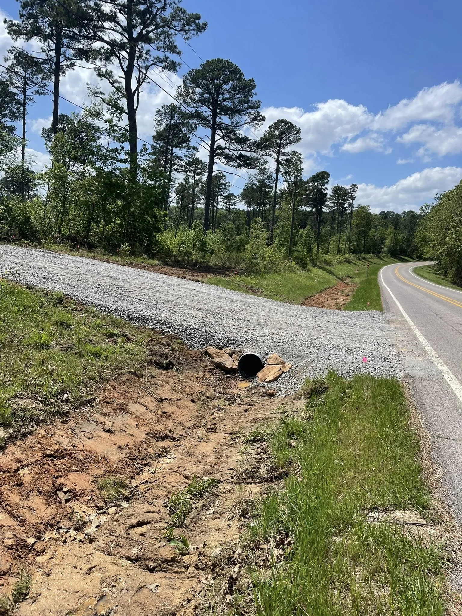 A rural scene with a winding road, trees, and a mix of clear sky and clouds, with some construction or maintenance work on the side of the road.