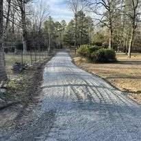 Gravel driveway lined with trees and shrubs in a rural setting.