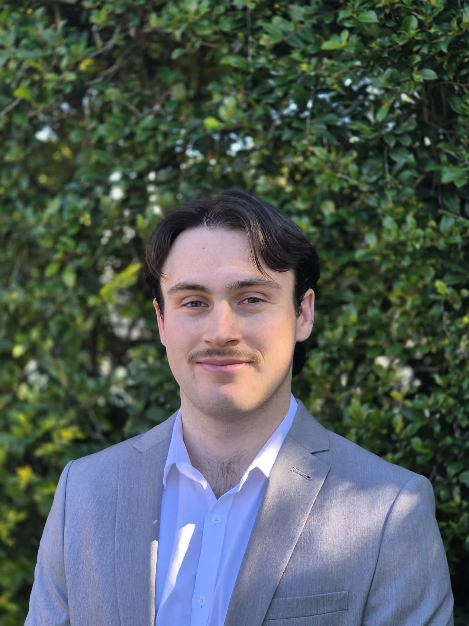 A portrait of a young man with dark hair, wearing a light gray suit and white dress shirt, standing outdoors in front of green bushes.