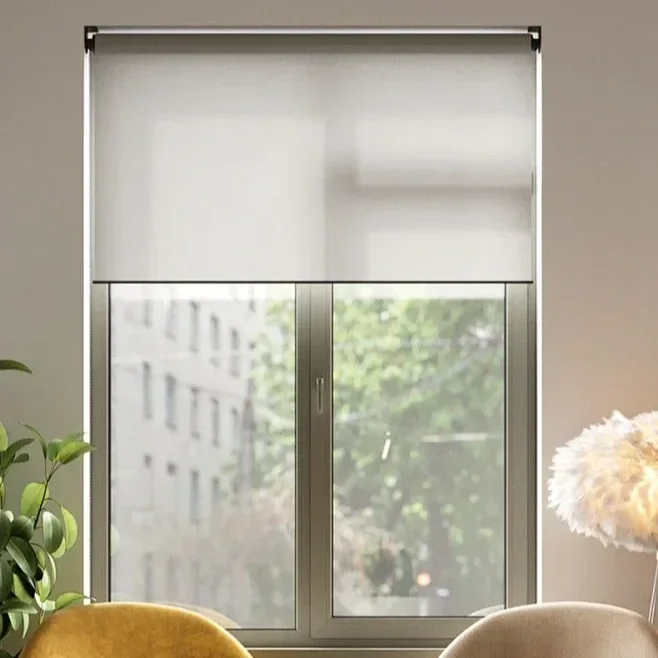 A window with a white roller blind, partially lowered, with a view of a building and green trees outside.