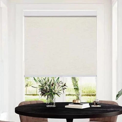 A window with a white roller shade partially rolled down, revealing a view of green trees outside. In front of the window is a round black table with a vase of flowers and a few books and candles.