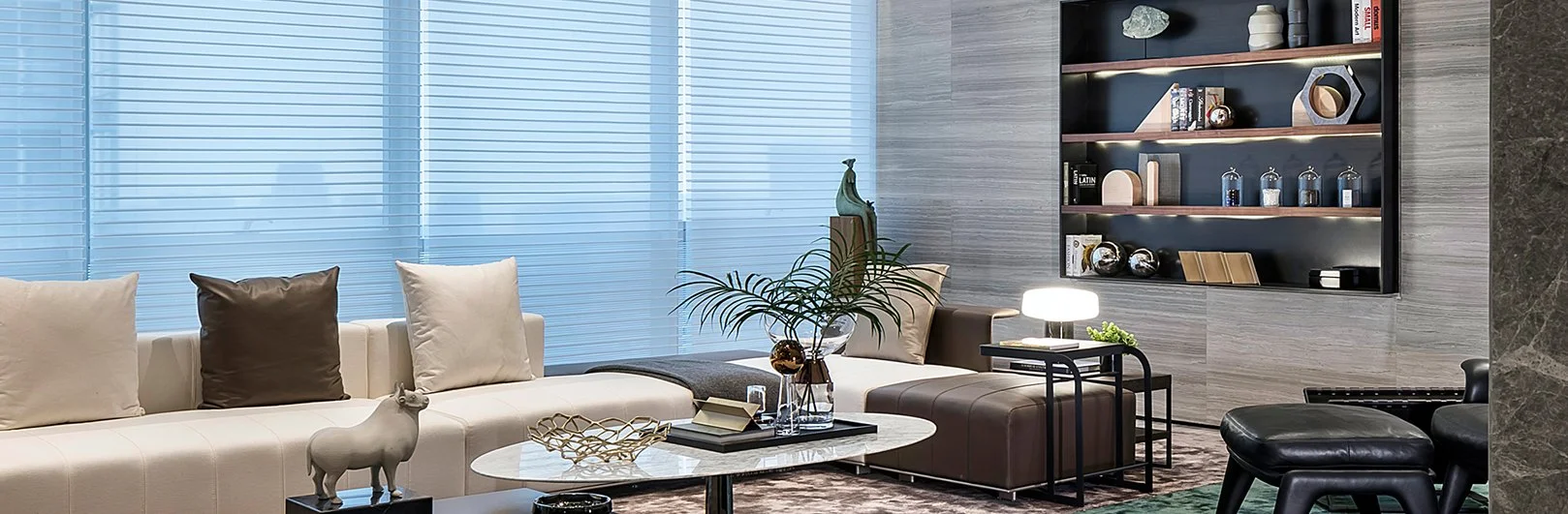 Modern living room with white sectional sofa, decorative pillows, and a coffee table with decorative items. Large windows with blue blinds, a side table with a lamp, and a black shelving unit with decorative objects on a textured wall.