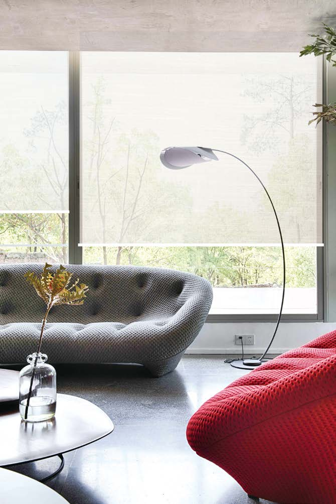 Modern living room with large window blinds, a gray tufted sofa, a red textured armchair, a white side table with a glass vase and plant, and a tall curved floor lamp.