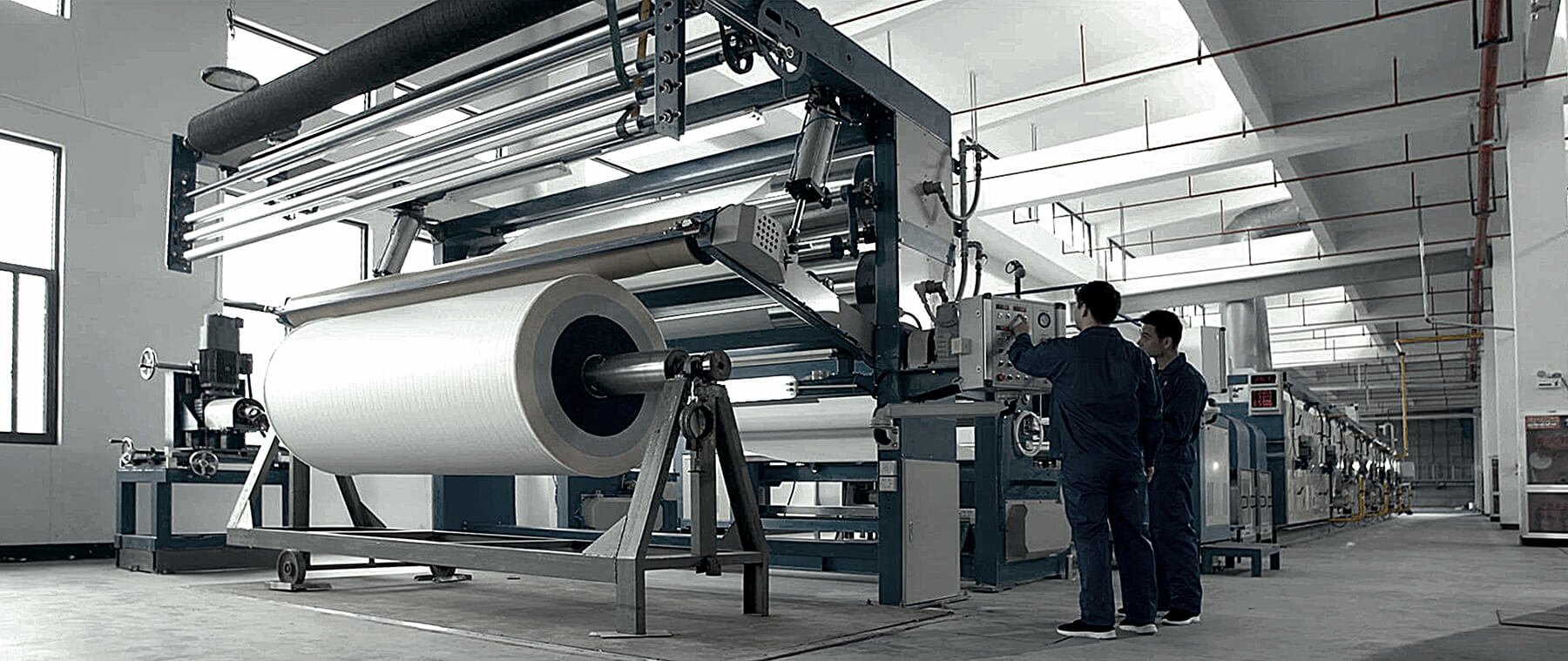Two workers operating a large industrial machine in a commercial textile manufacturing facility, processing a wide roll of fabric or paper.