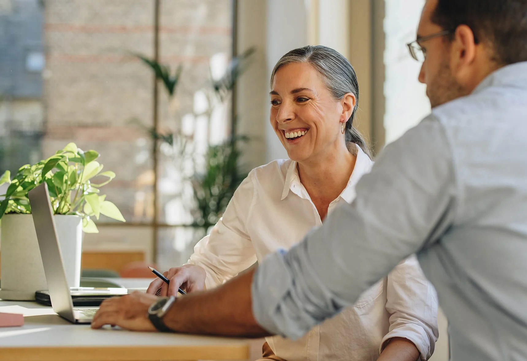 Two people, a woman and a man, sitting at a desk having a discussion, both smiling, with a laptop and a plant in the foreground. they are discussing career growth and preparing your CV for interviews.