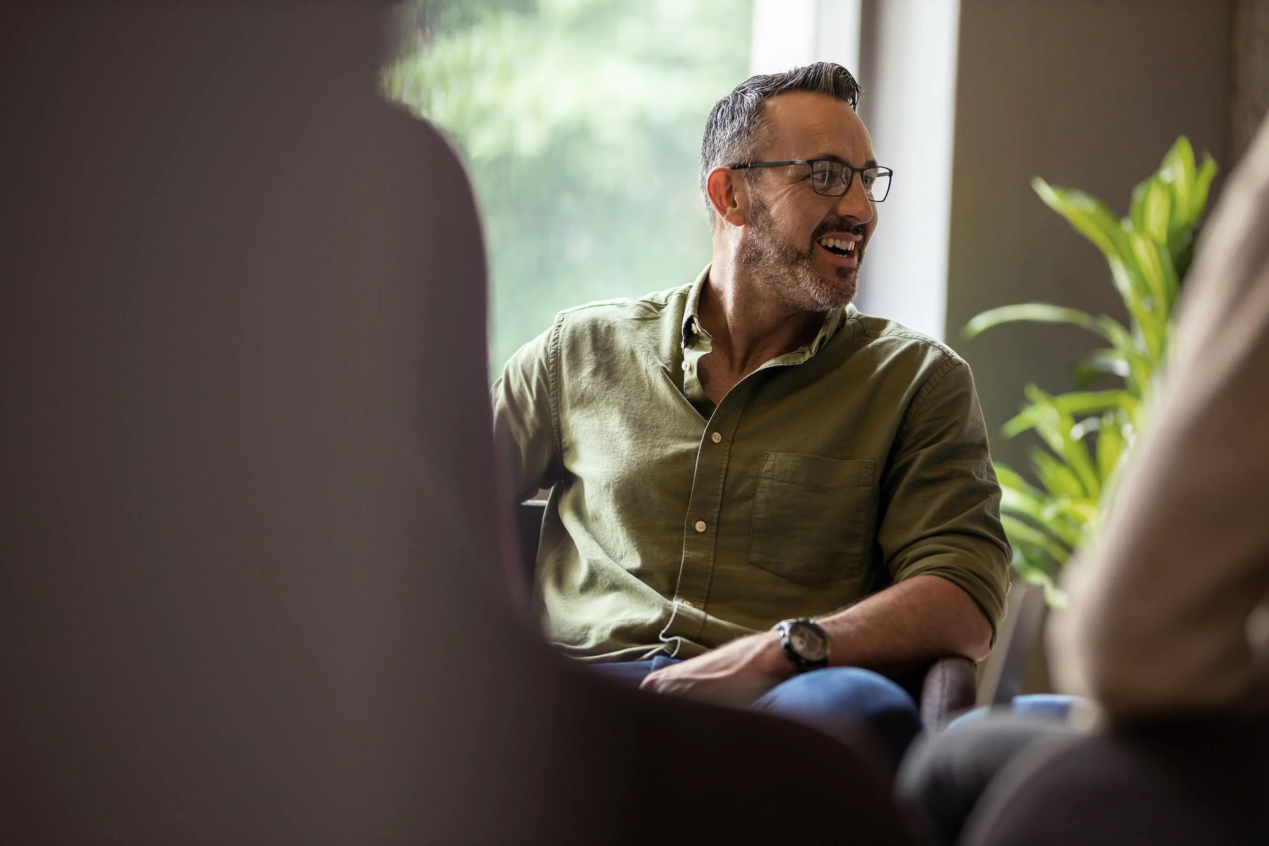 A man with gray hair, beard, glasses, talking during a meeting. He is enjoying getting job satisfaction through career counselling.