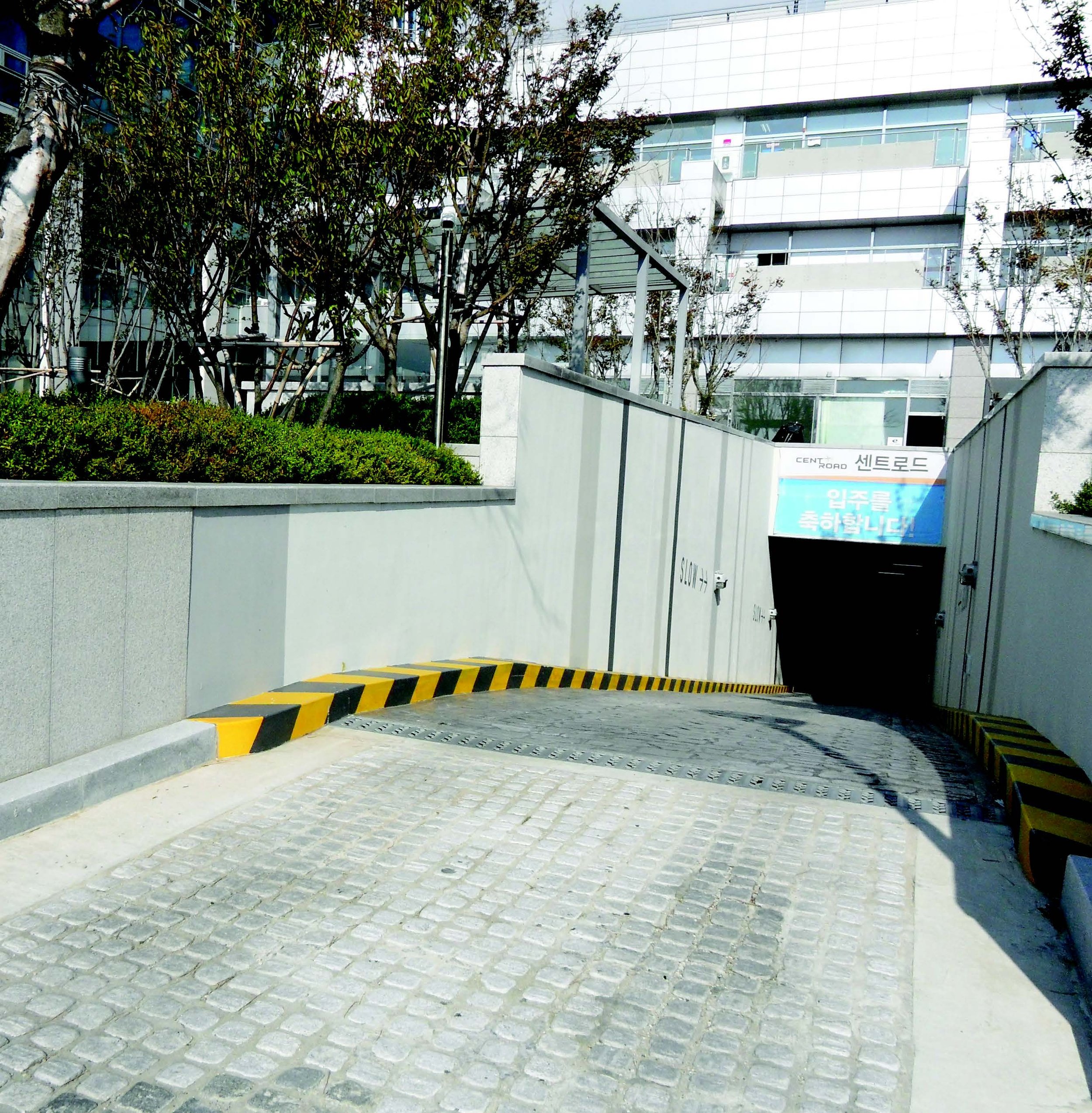 An outdoor scene with a modern building in the background surrounded by trees and bushes. The building has multiple floors with glass windows and balconies. There is a sign with Korean characters, and a parking area or driveway with a protective barrier painted in yellow and black stripes.