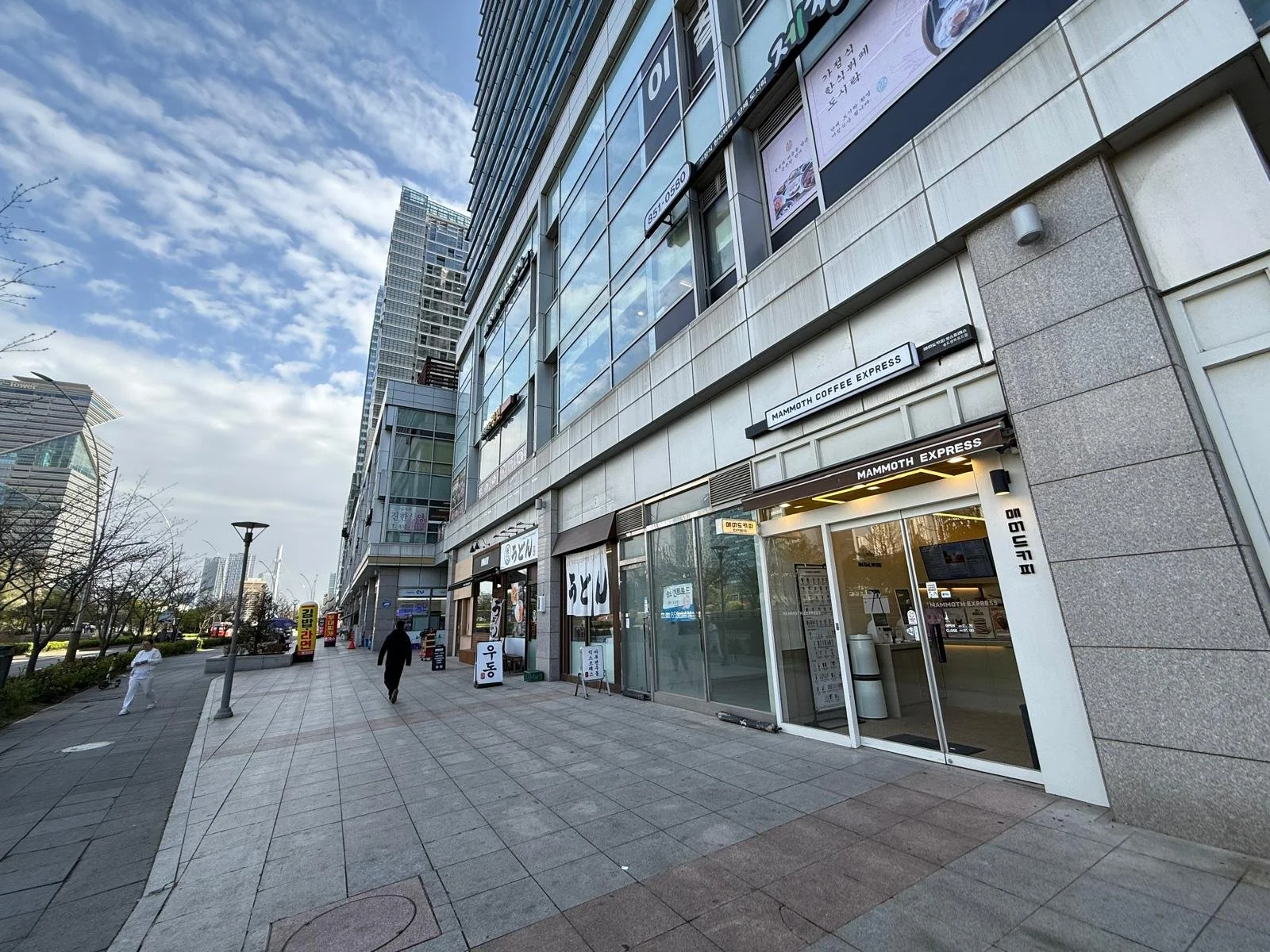 City sidewalk scene with a modern building, people walking, and a mammoth coffee shop visible in the foreground, during daytime with partly cloudy sky.