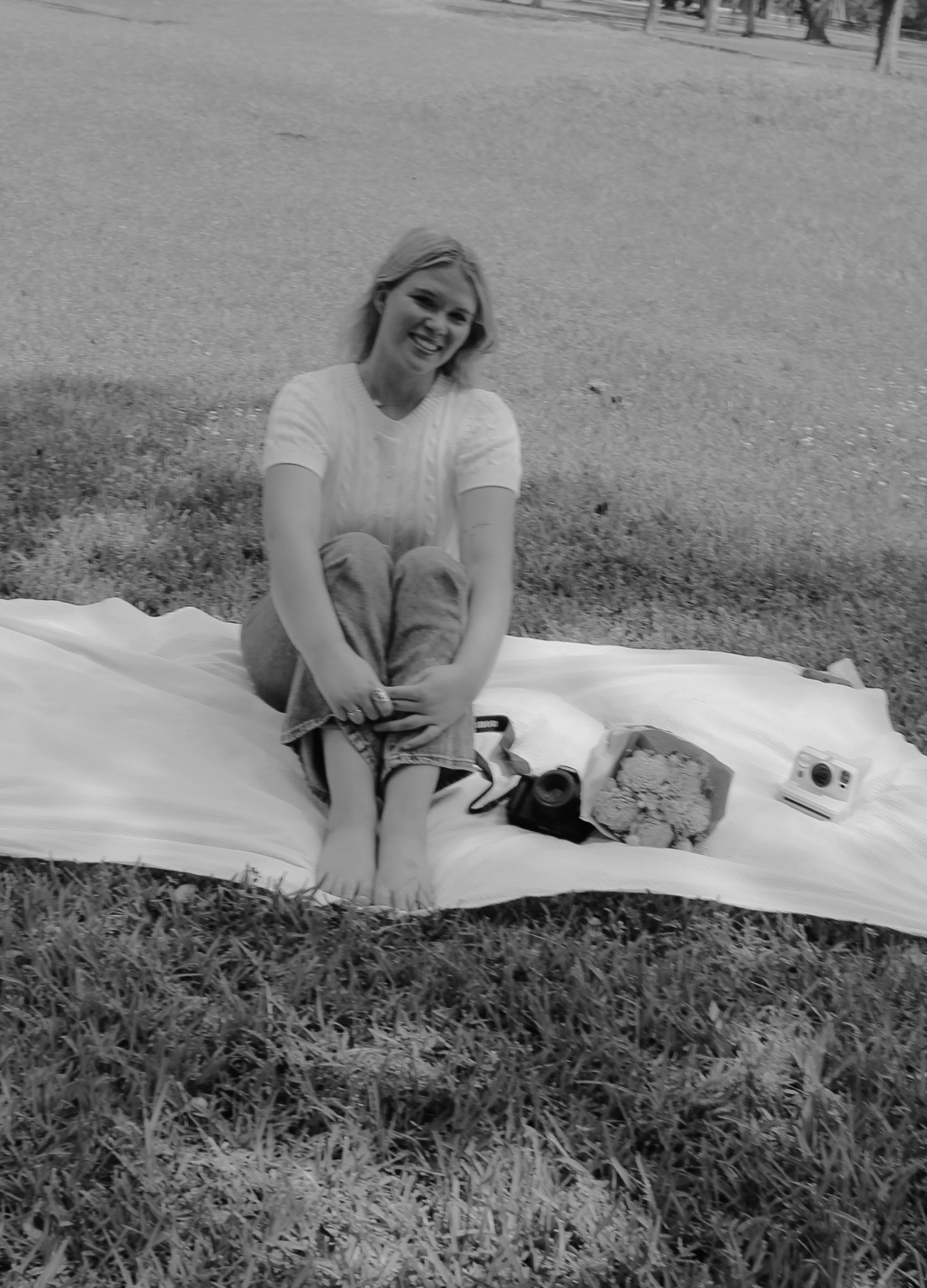 A woman with short hair and a light-colored shirt sitting on a blanket in a park with grass and trees in the background. She is smiling and has her arms wrapped around her knees. There are items on the blanket, including a container of food, a toy camera, and some snacks.