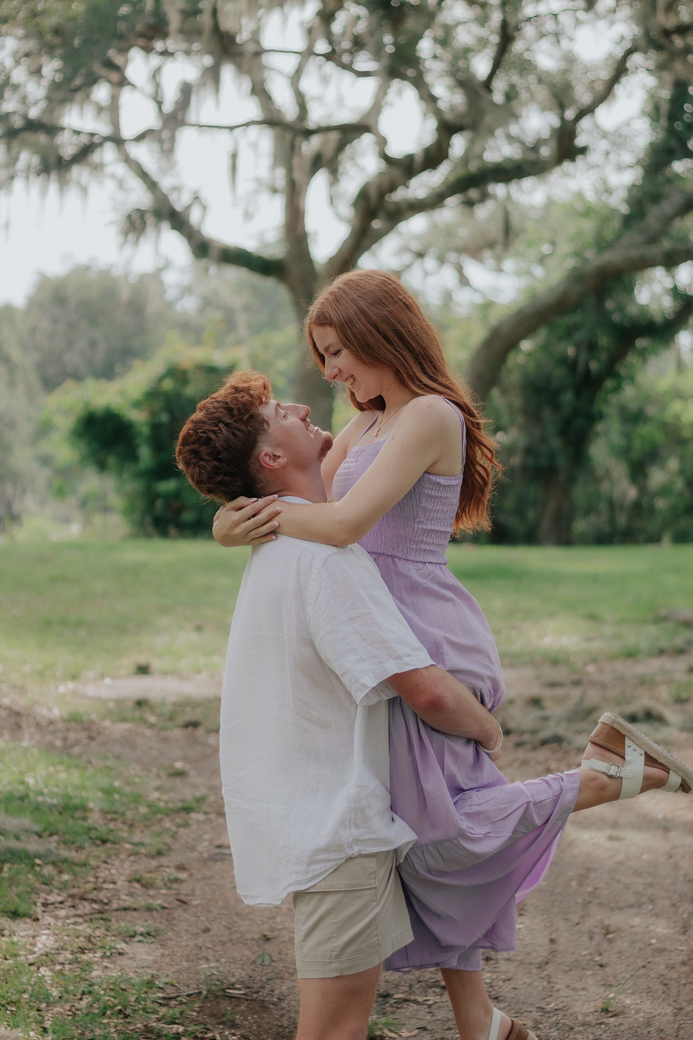A young man is holding a young woman in his arms and kissing her on the forehead outdoors in a park with trees in the background. The woman is smiling and looking down at the man, and they both appear happy.