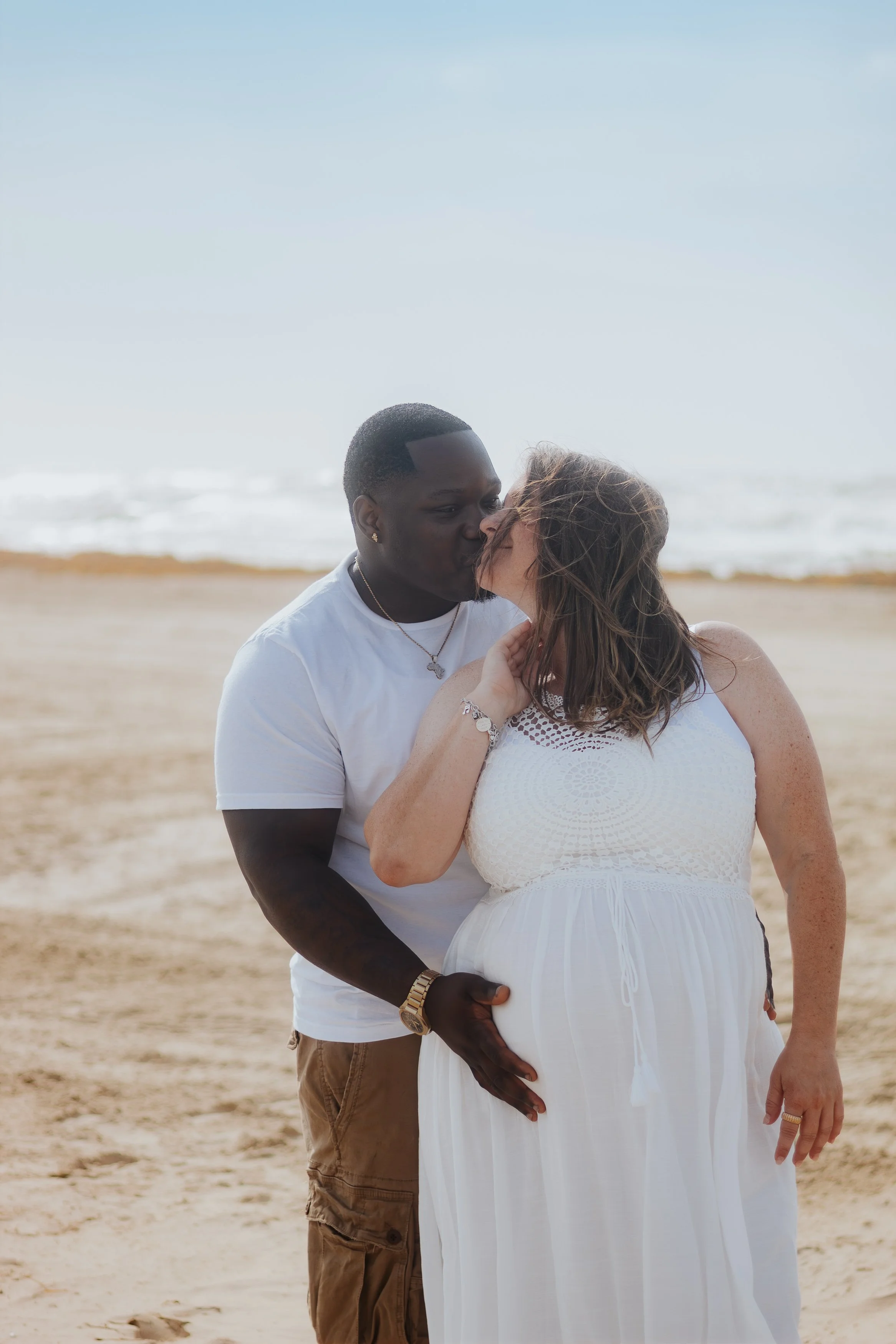 A couple kissing on the beach, with the man gently touching the woman's pregnant belly.