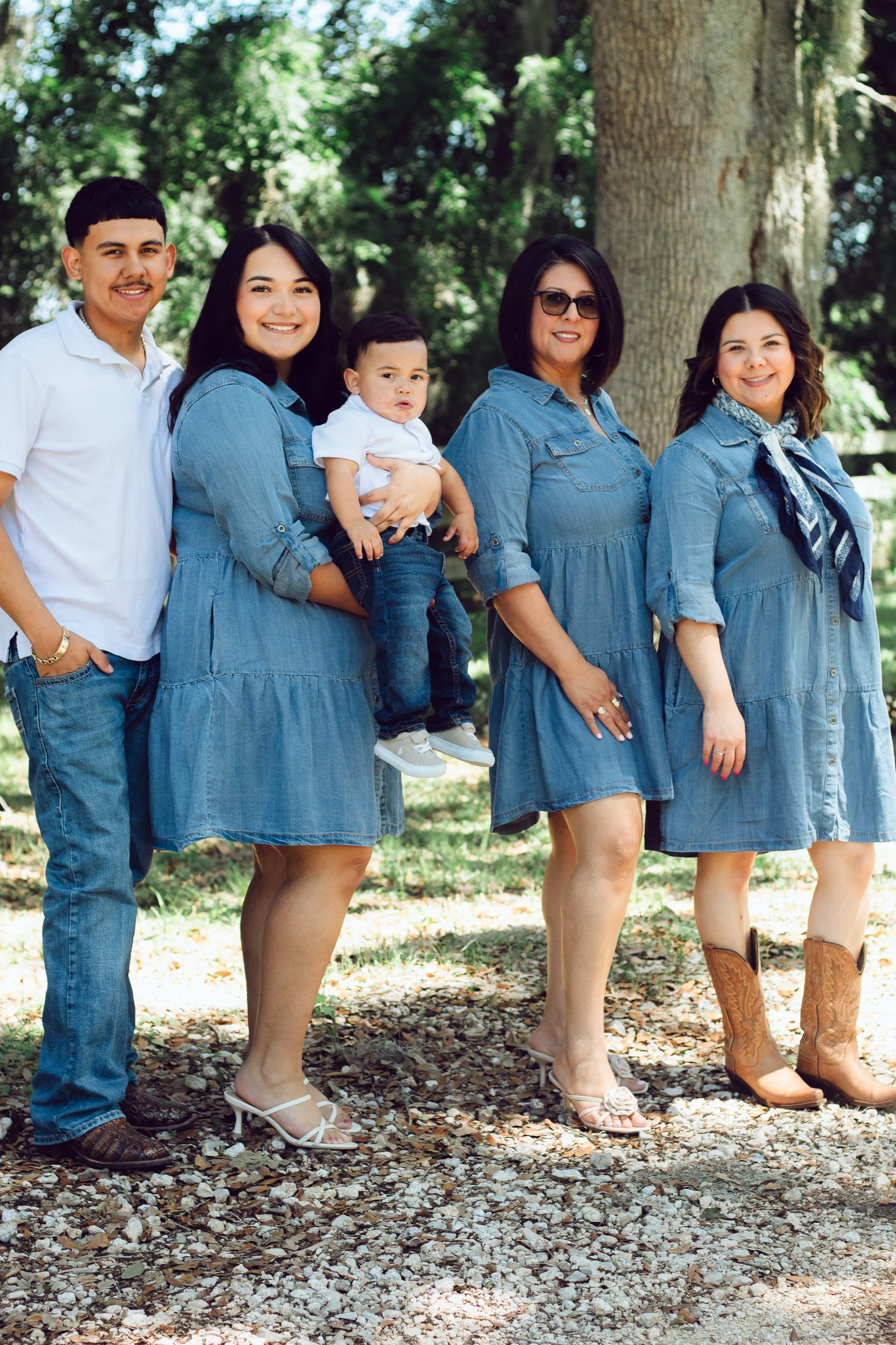 Group of five women and one man standing outdoors, wearing denim dresses and casual footwear, with a background of trees and rocky ground.