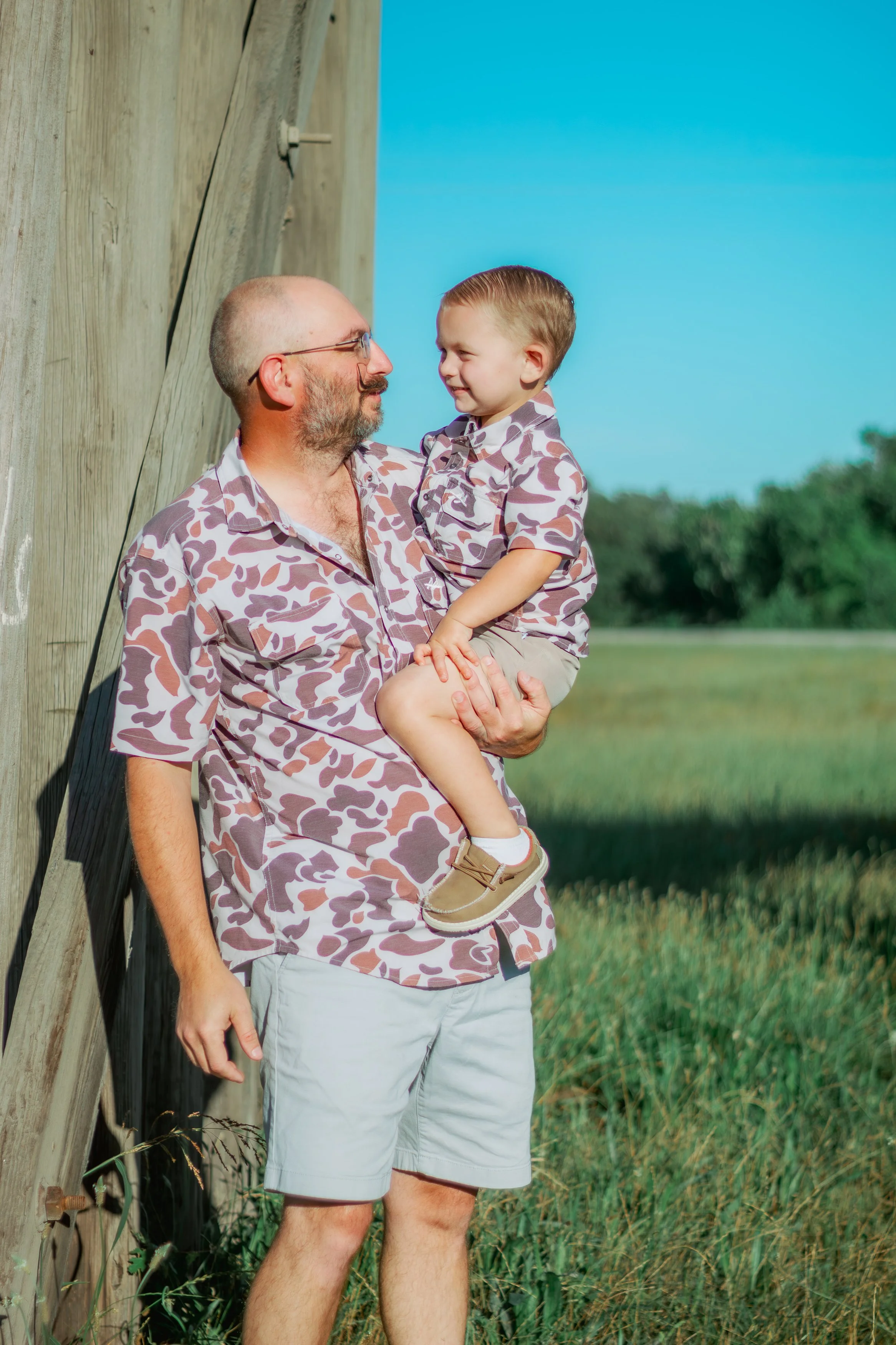 A man holding a young boy outdoors near a wooden structure, with a field and blue sky in the background. Both are wearing matching camouflage shirts.