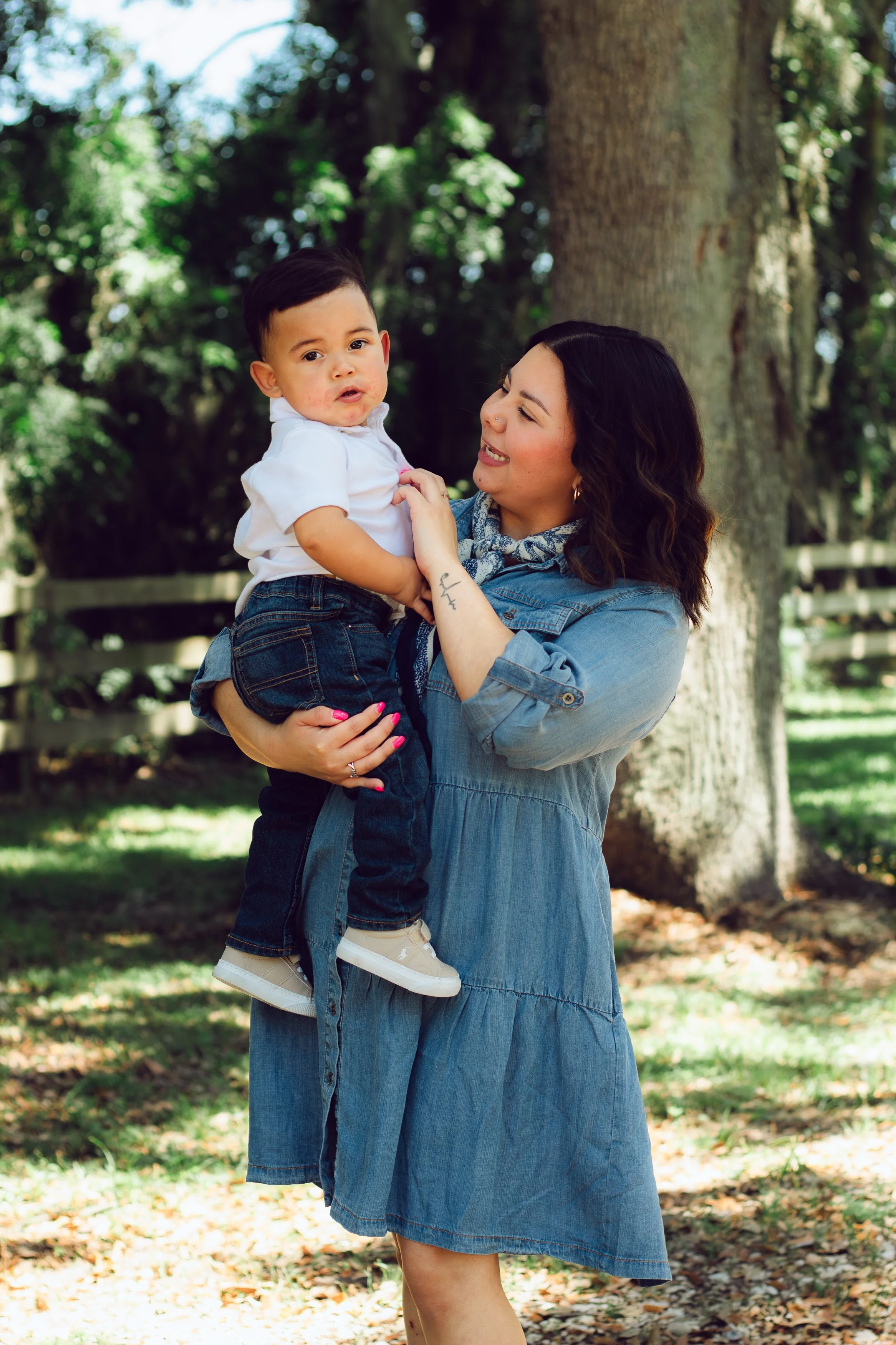A woman holding a young boy outdoors in front of a large tree, with a wooden fence and greenery in the background.