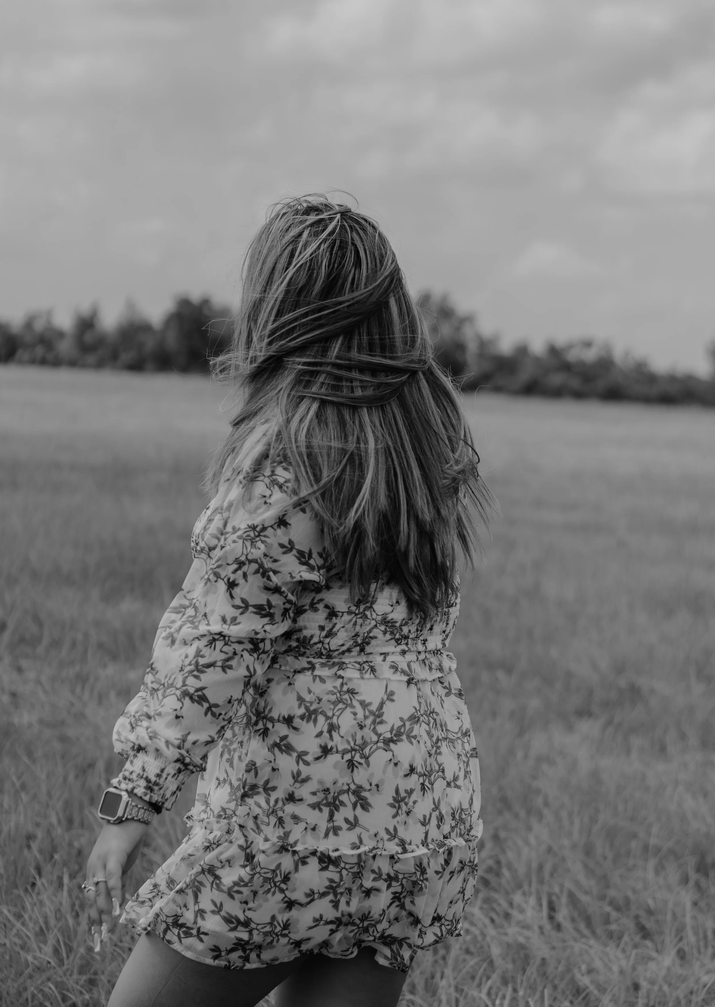 A woman with long hair, wearing a floral dress and smartwatch, standing outdoors in a field with trees in the background, black and white photograph.