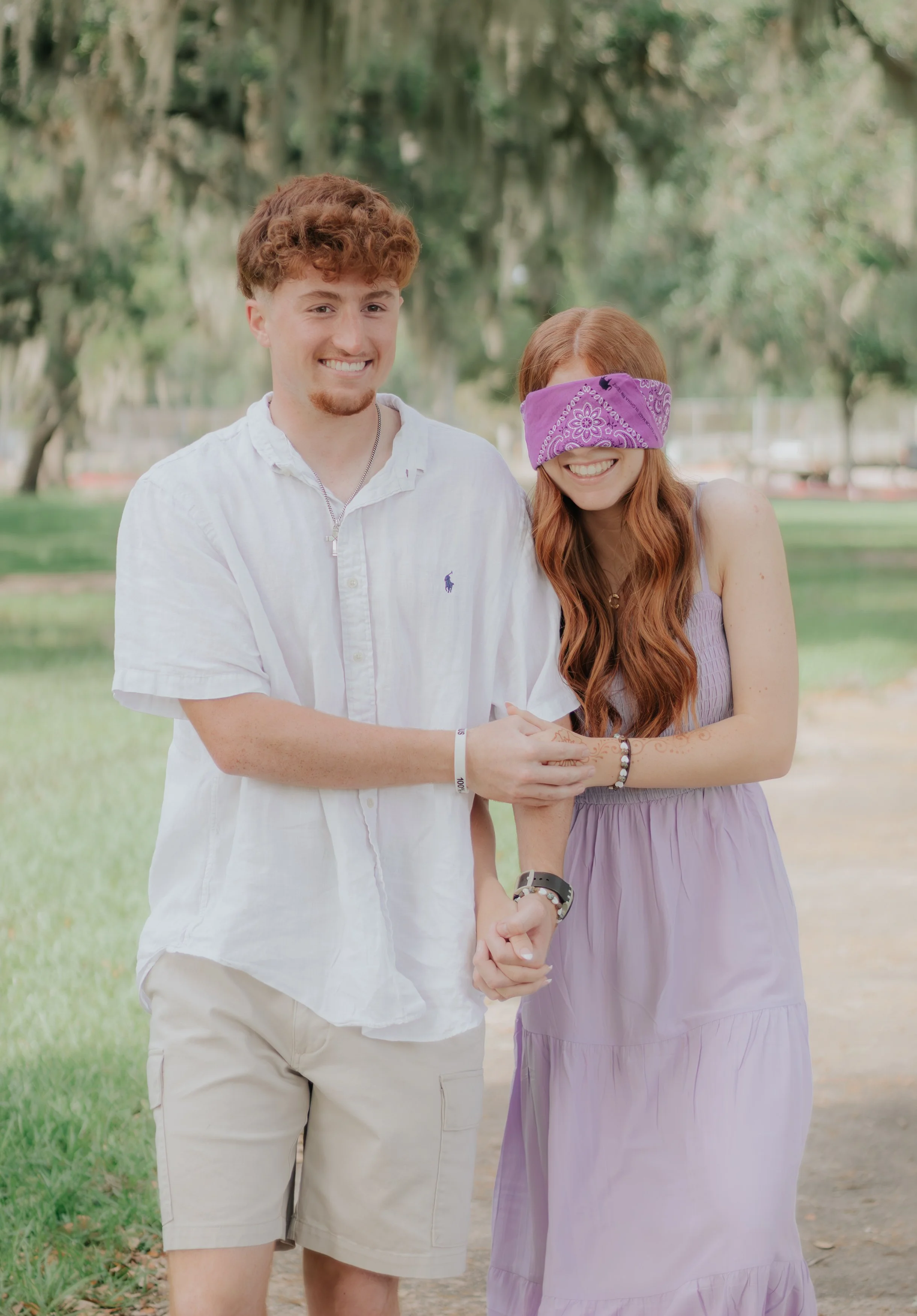 A young man and woman standing outdoors in a park with trees in the background. The woman is blindfolded with a purple bandana and smiling, while the man beside her is holding her hand and smiling.