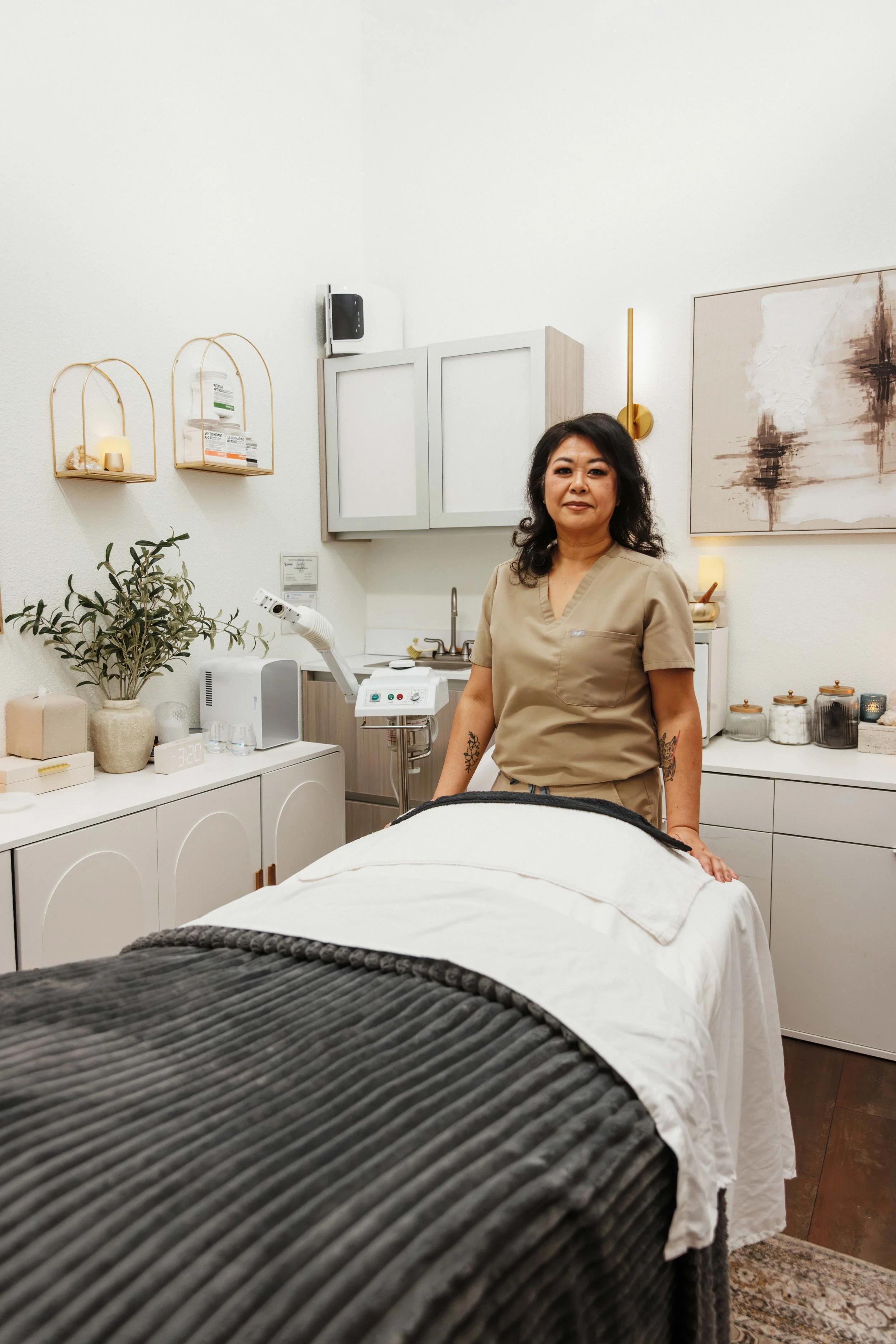 A woman in beige scrubs standing next to a massage table in a spa or treatment room with white walls, decorative shelves, and various jars and decor.