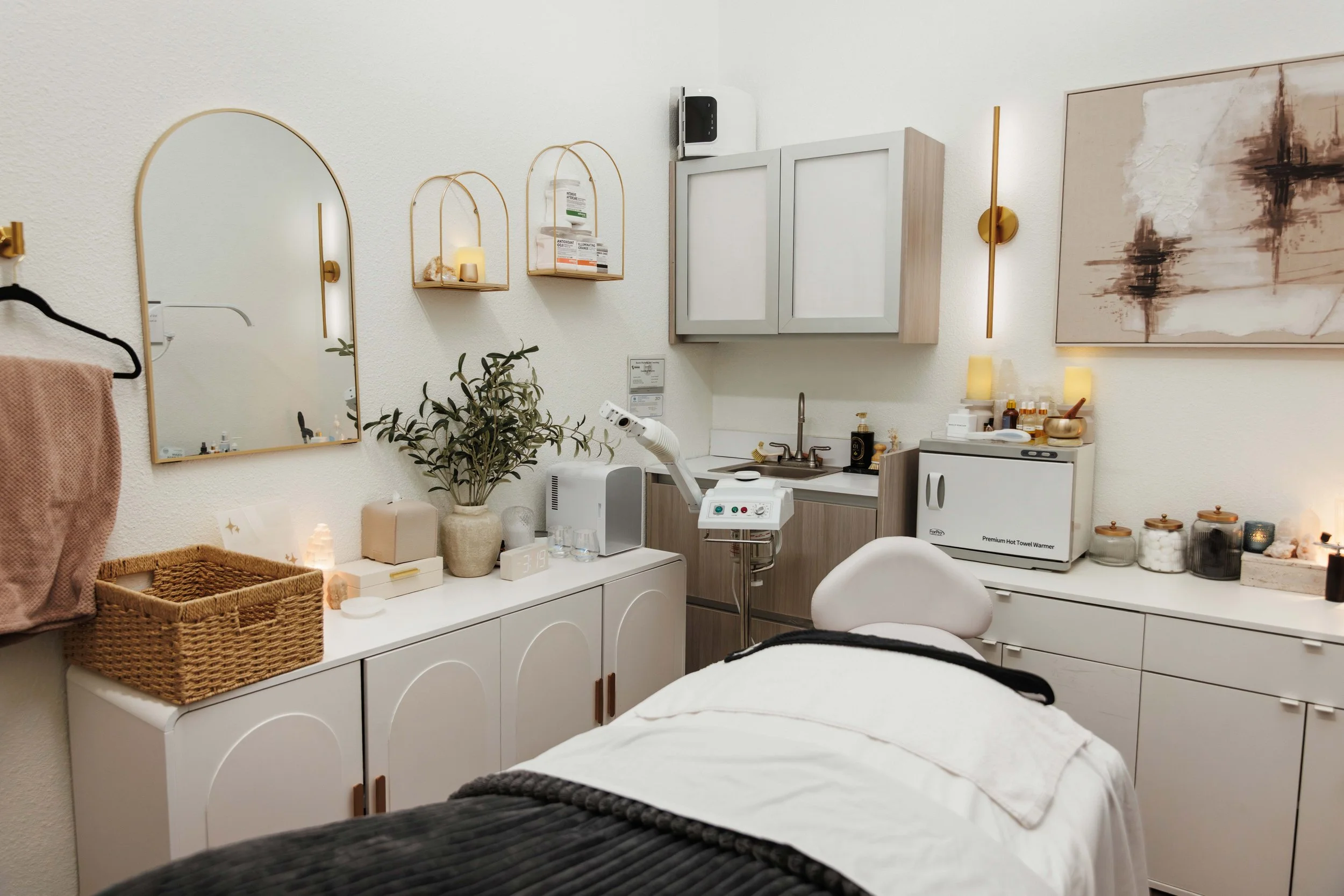 Interior of massage or wellness room with massage table, botanical decor, mirrors, shelves with skincare products, and wellness equipment.