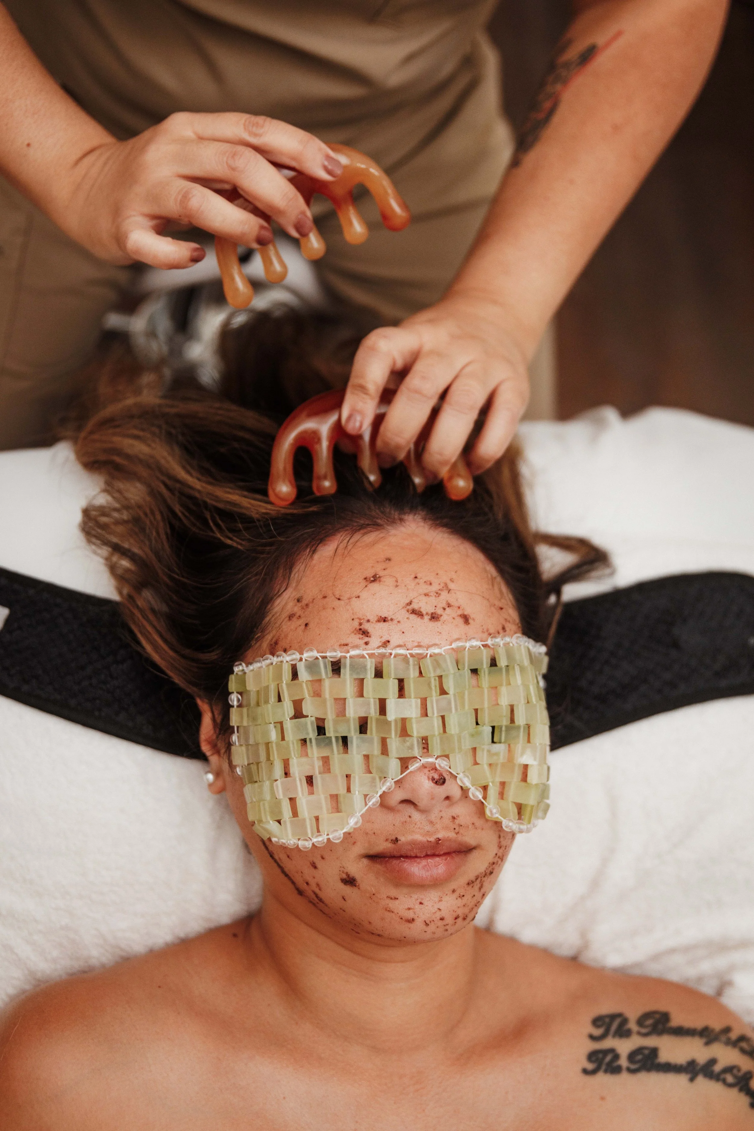 A woman lying on a treatment table with facial microblading markings, wearing a woven eye mask, and another person applying facial wax with fingers and waxing strips on her face.