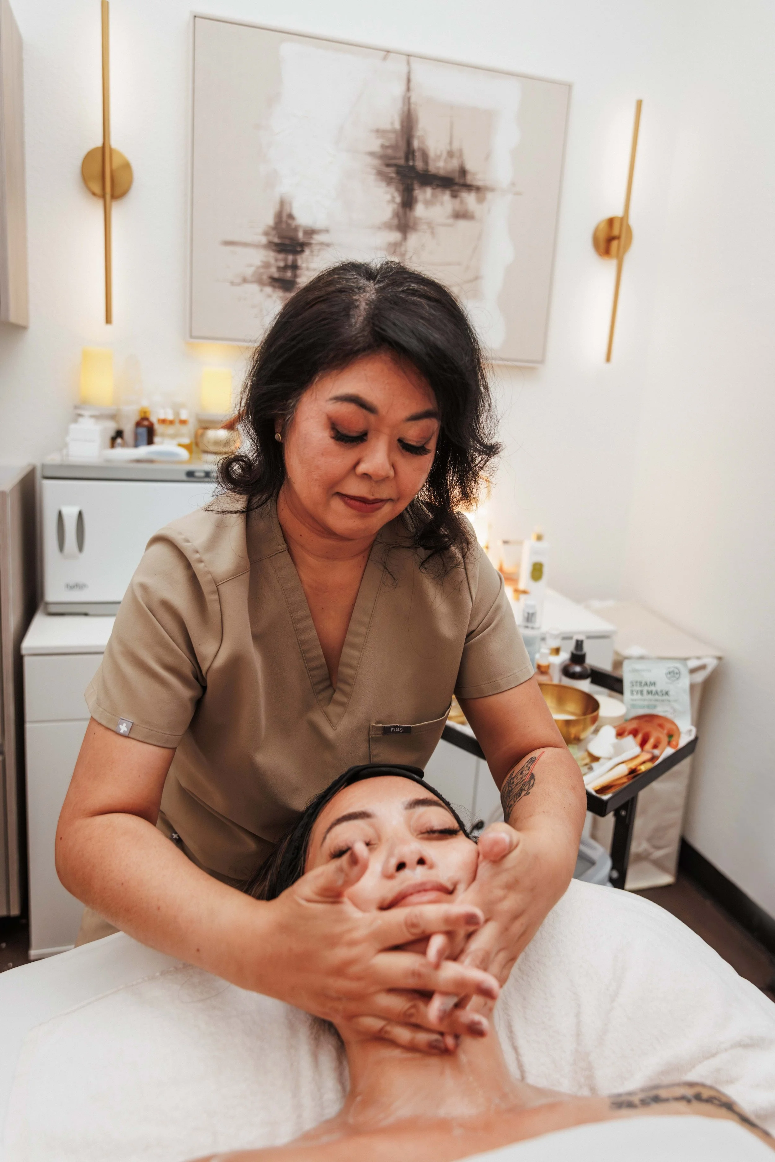 A woman receiving a facial massage or treatment from a professional at a spa or clinic, lying on a treatment bed with her eyes closed.