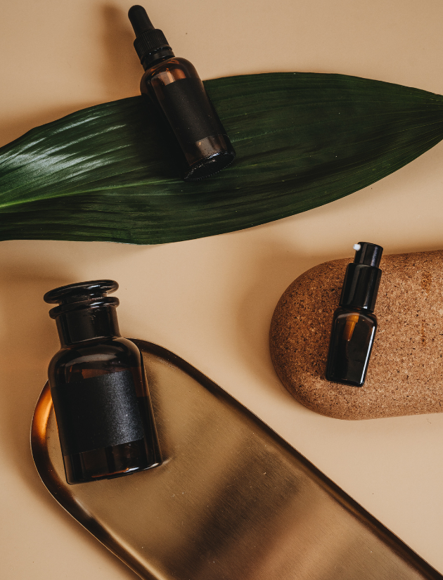 Three small dark amber glass bottles with black caps, a large green leaf, a brown stone, and a metallic tray on a beige surface.