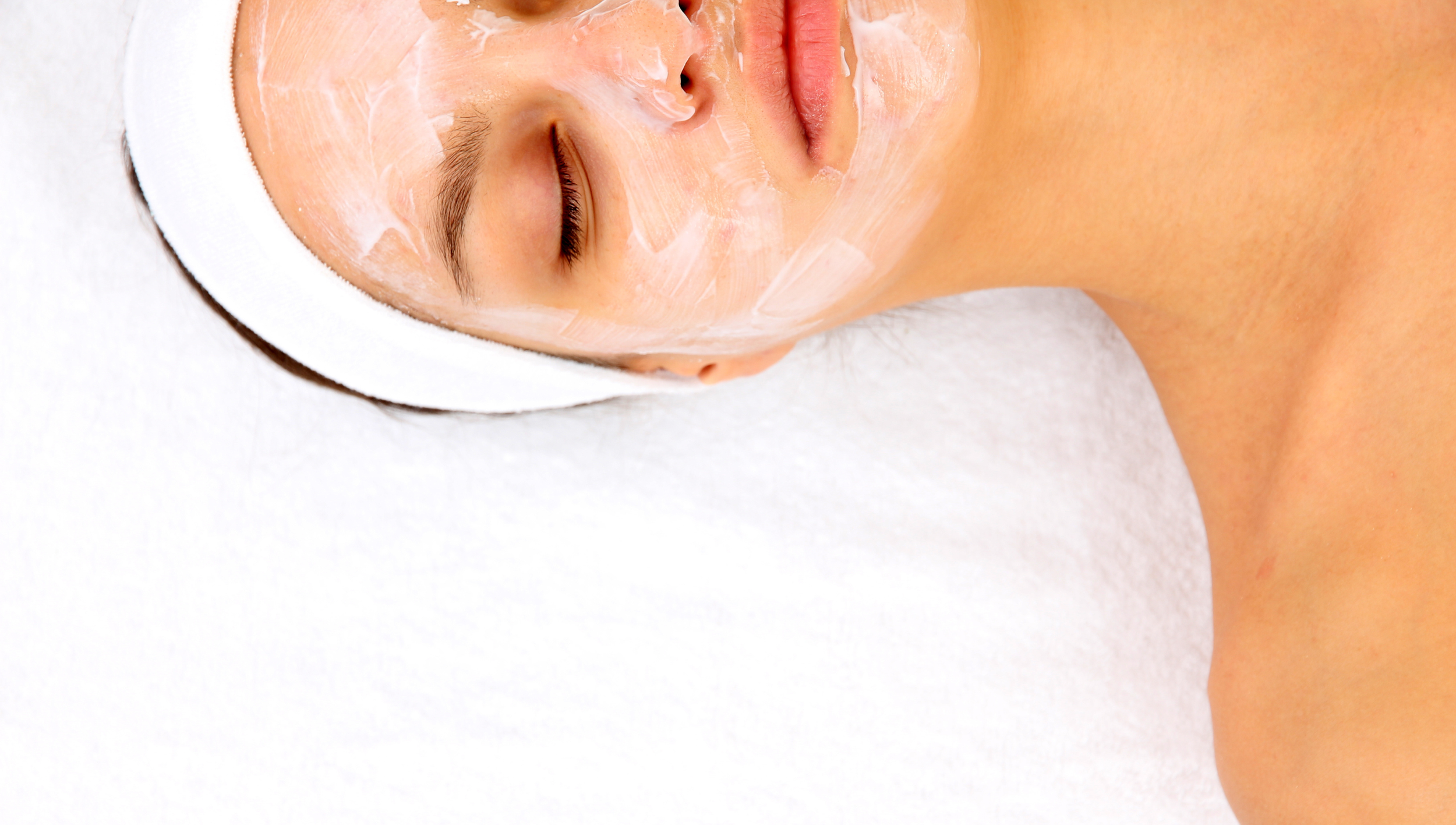 A woman lying down with her eyes closed, receiving a facial treatment; her face is covered with a white cream or mask, wearing a white headband, on a white surface.