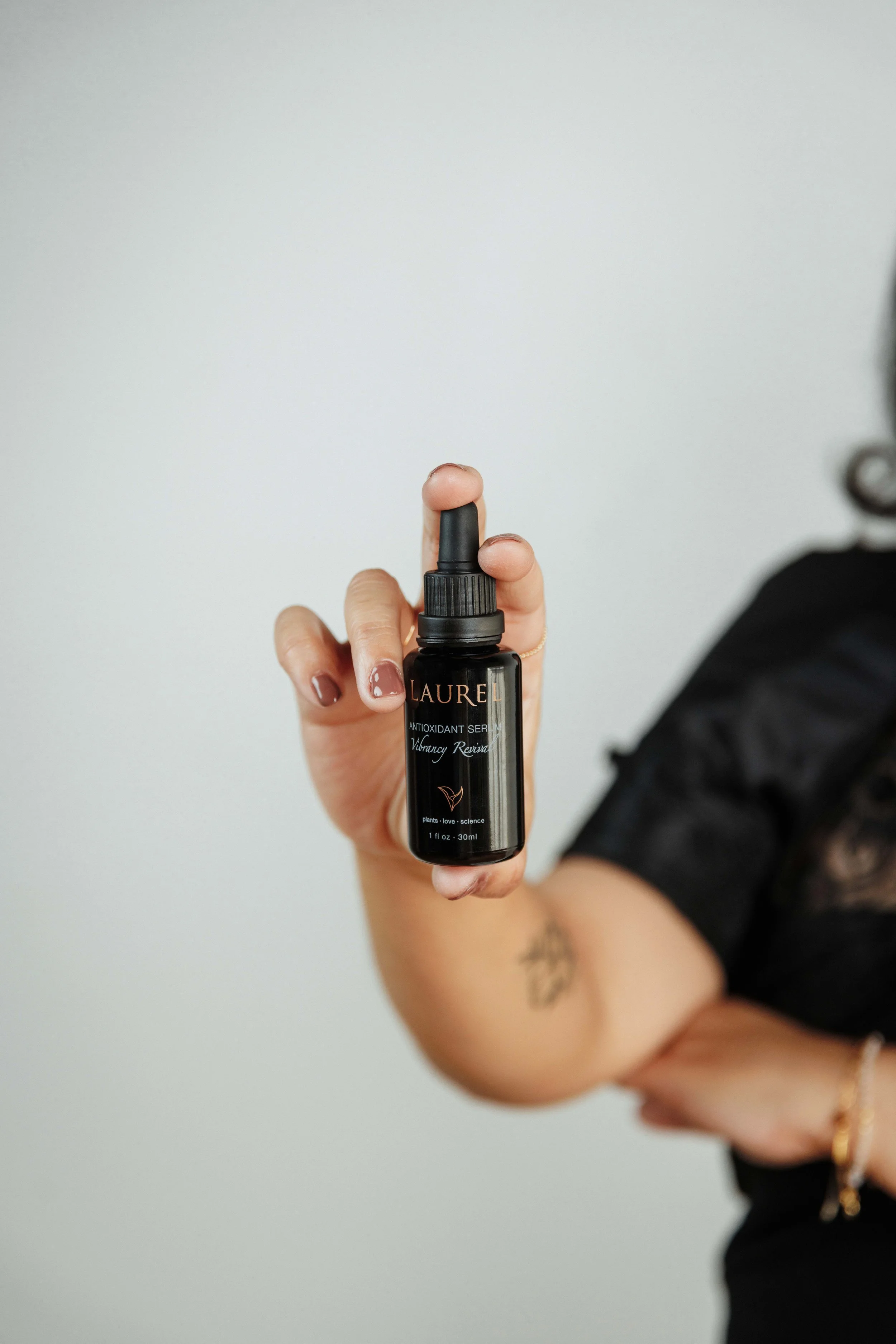 Person holding a small black bottle labeled 'Laurel Antioxidant Serum' with a dropper cap, partially visible arm and torso, against a plain light background.