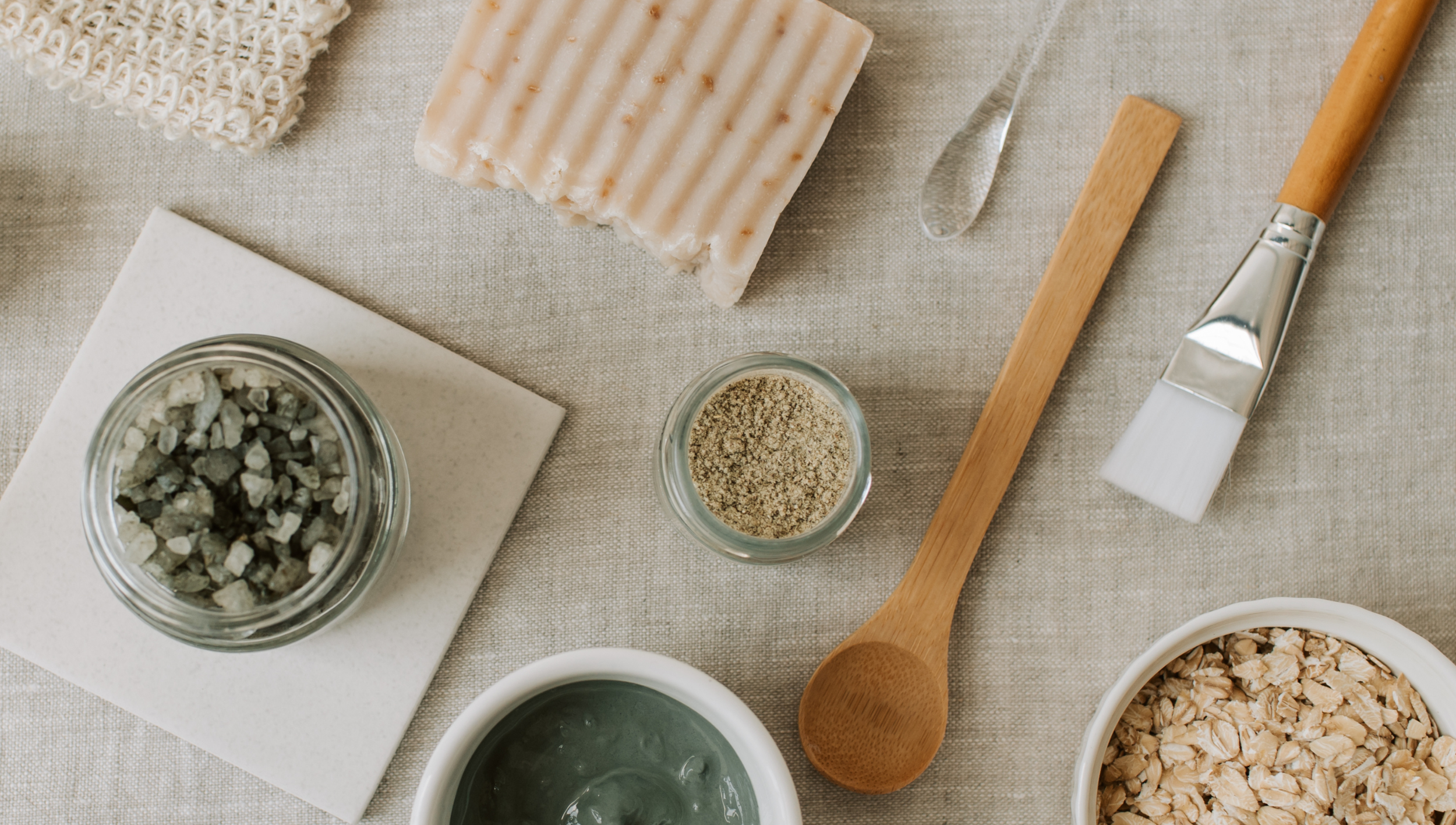 Natural soap, ceramic bowl with gray-green clay, jar of gray and white stones, small jar of ground herbs, oatmeal in a white bowl, and various soap making tools on a beige fabric surface.