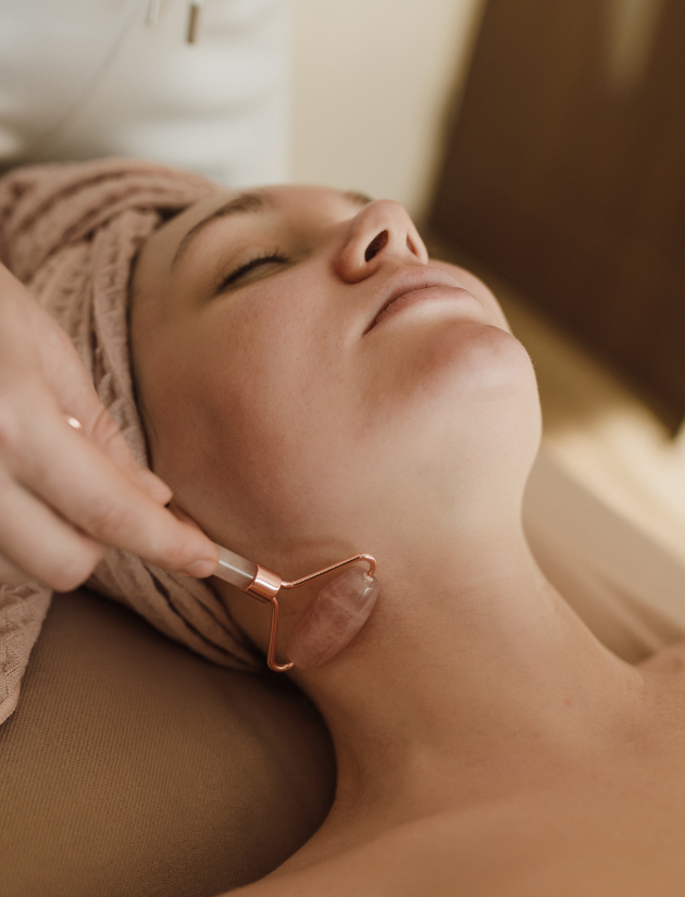 A woman lying down with her eyes closed, receiving a facial or skincare treatment using a jade roller on her neck.