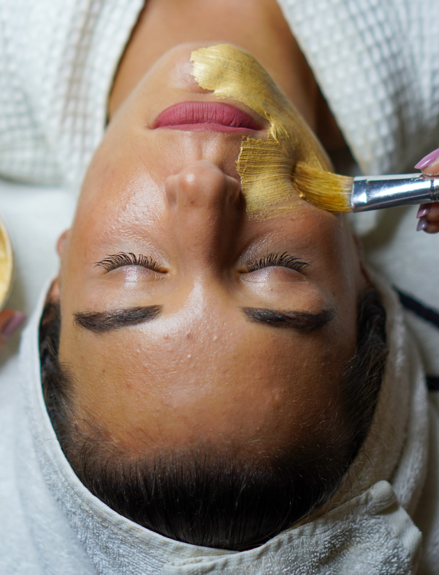 A woman receiving a facial treatment with gold-colored mask being applied to her face by a professional.
