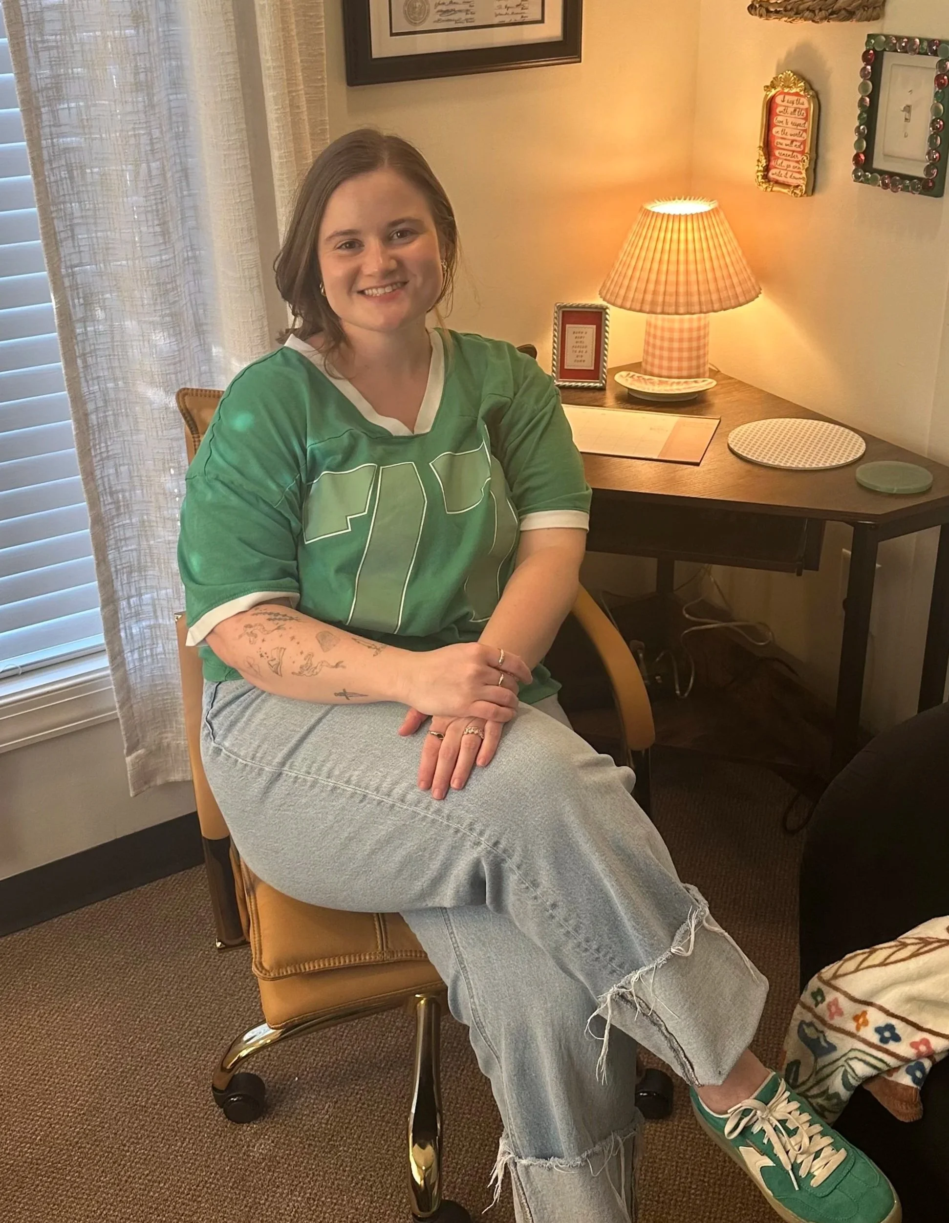 Young woman sitting on a tan office chair with her right arm resting on her lap, smiling at the camera. She is wearing a green sports jersey, ripped light blue jeans, and green sneakers. She has shoulder-length brown hair and tattoos on her right forearm. The background features a desk with a table lamp, picture frames, and decorative items, beside a window with white blinds and sheer curtains.