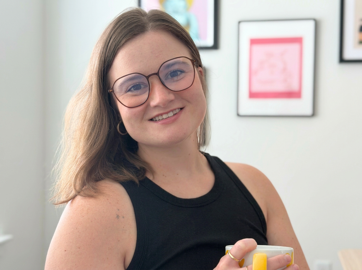 A young woman with shoulder-length brown hair, wearing glasses and a black sleeveless top, smiling while holding a white mug with yellow accents.