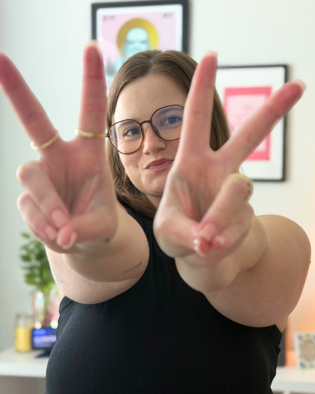 A woman with glasses making peace signs with both hands, smiling, with framed artwork and a plant in the background.