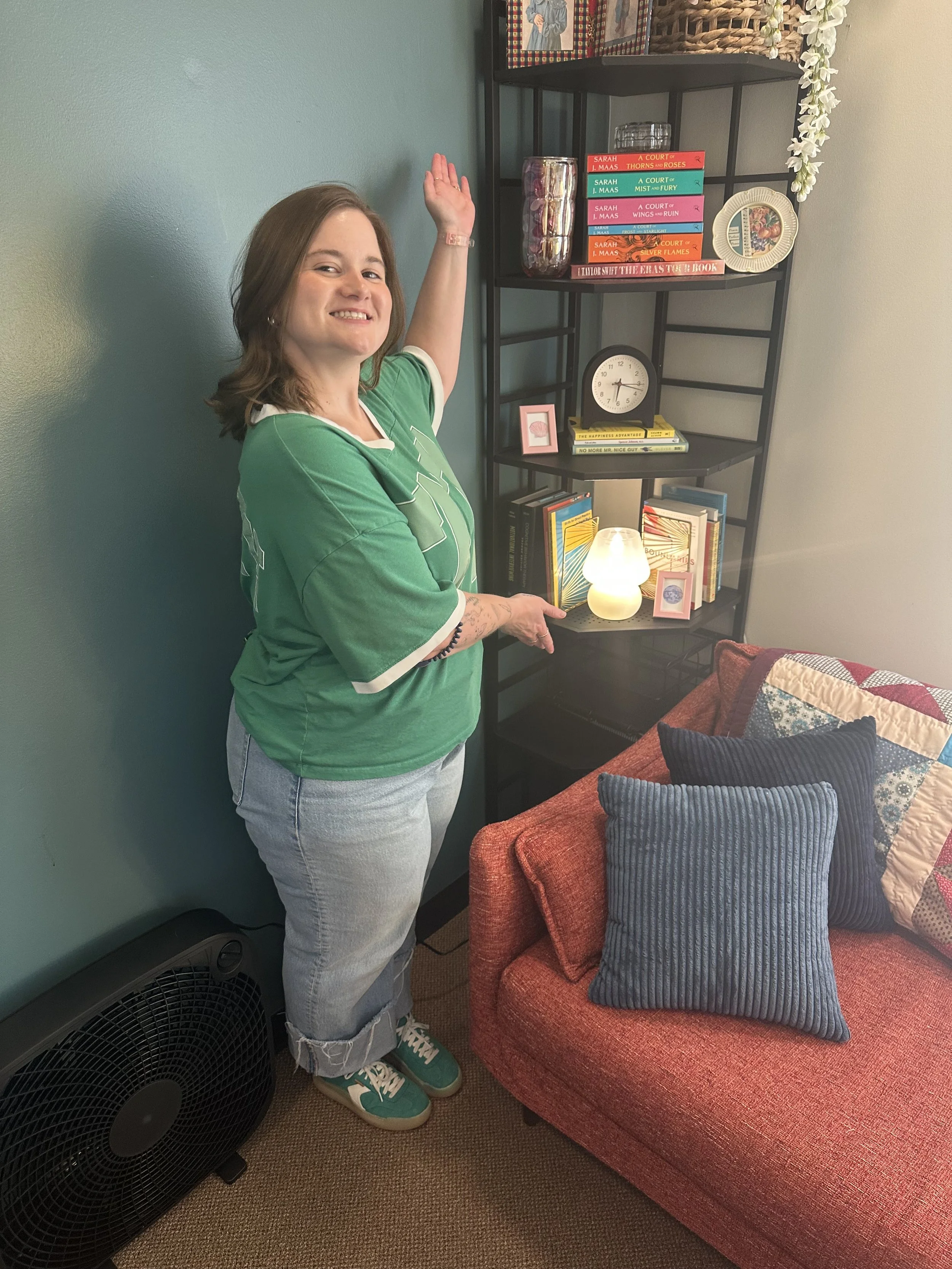 A woman with shoulder-length hair, wearing a green sports jersey, light jeans, and teal sneakers, stands next to a red fabric sofa with two gray striped pillows. She is, inside a room with a black fan on the floor next to the sofa, and a black shelving unit behind her with books, a clock, and decorative items. She is smiling and has one hand raised while the other points towards the shelf.