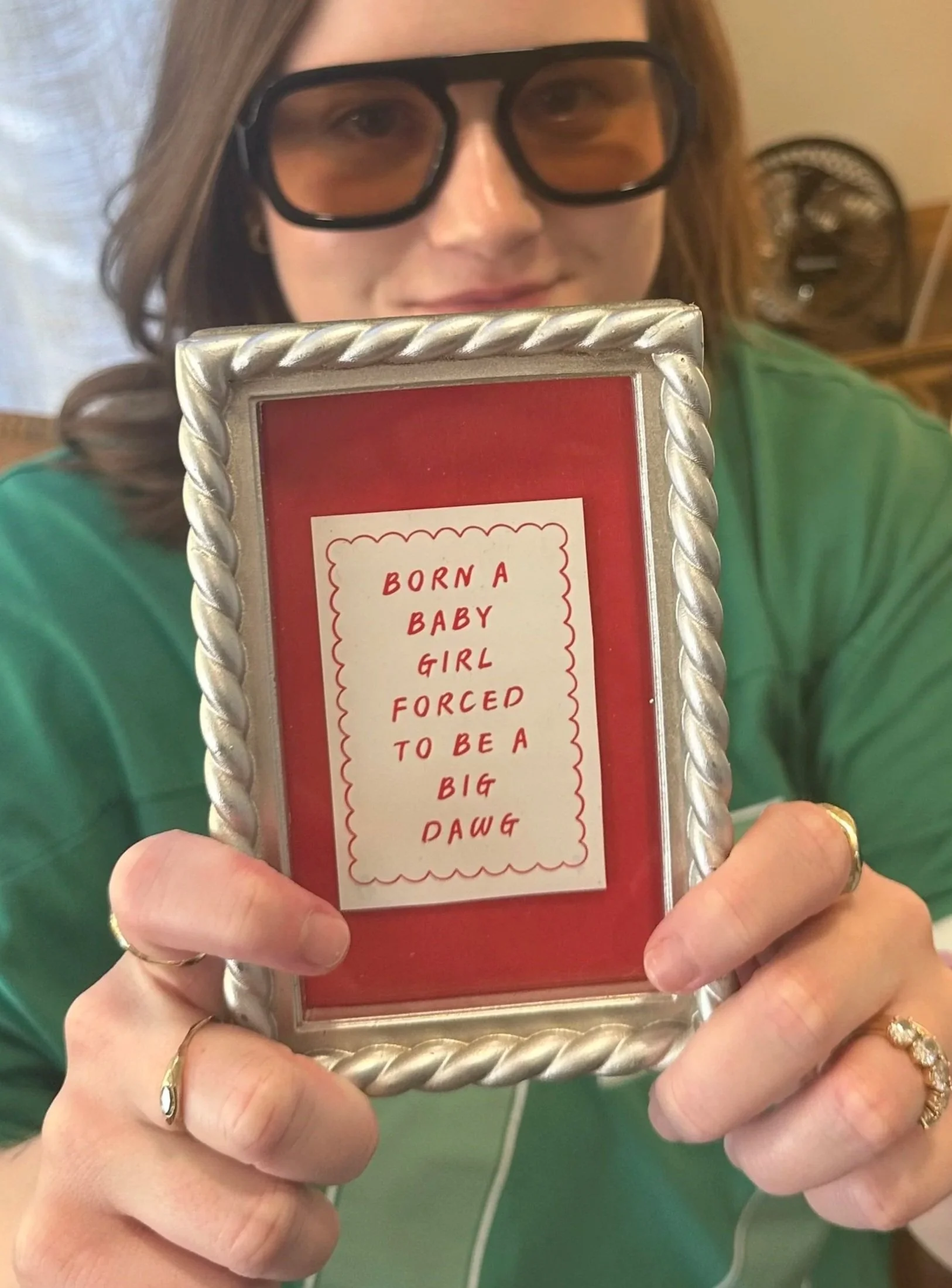 A person with light skin and wearing glasses is holding a red framed sign with a message. The person is wearing a green shirt and gold rings on their fingers. The background includes a fan and some wooden furniture.