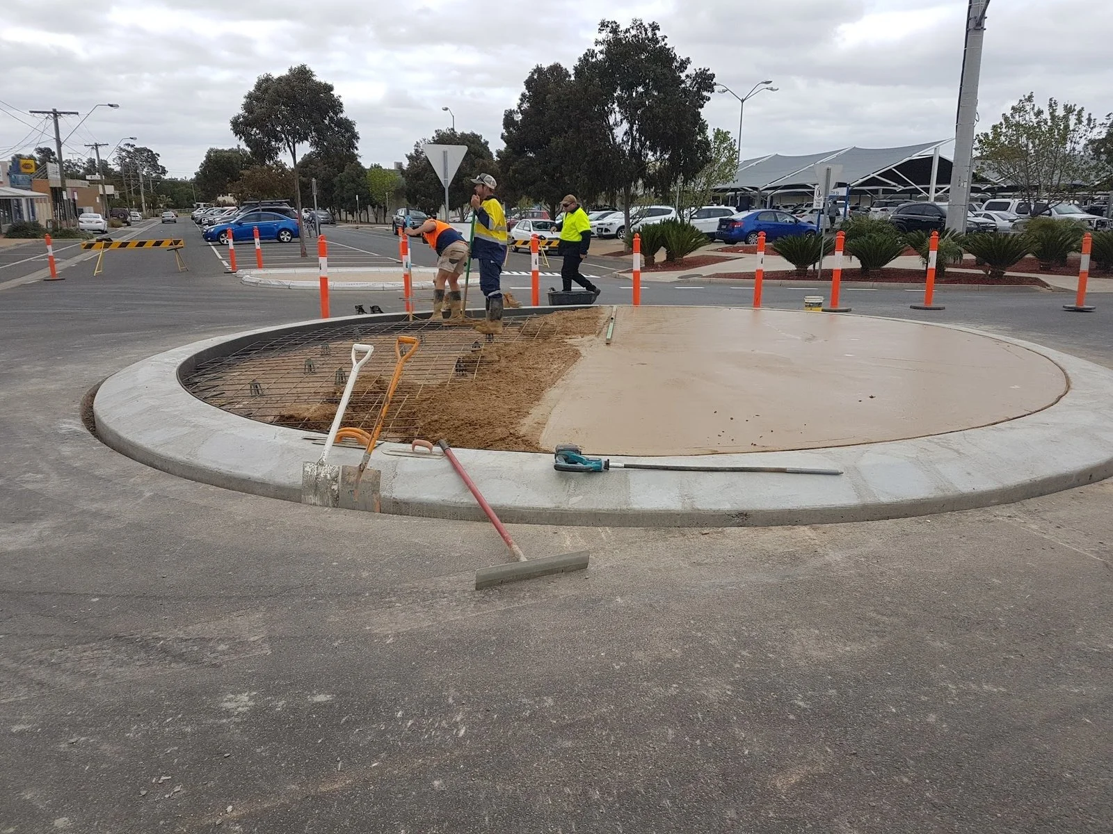 Construction workers installing a circular concrete feature in a parking lot near a street, with construction tools and orange safety cones around.
