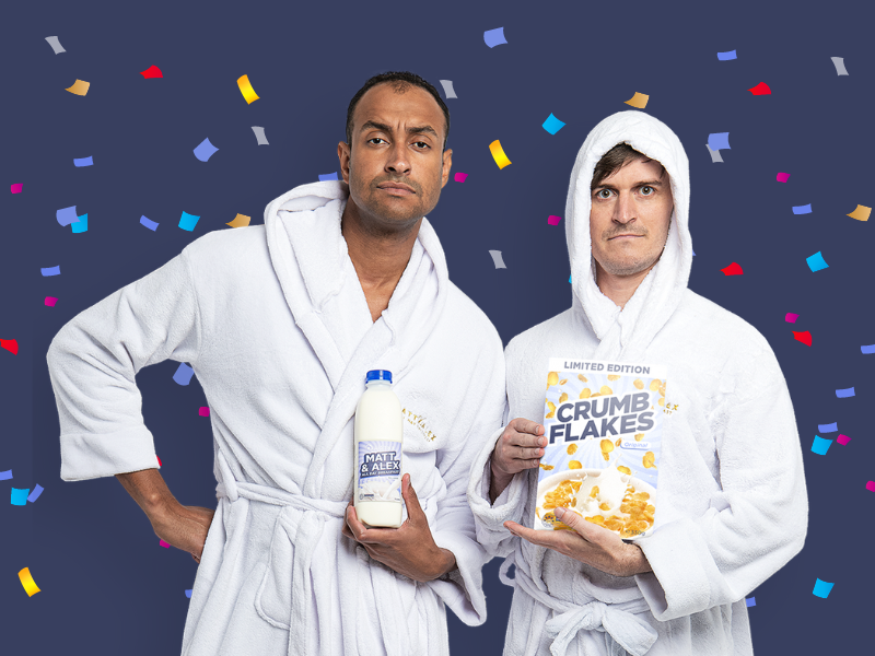 Two men in white bathrobes standing in front of a dark background with colorful confetti. One man holds a bottle of milk, the other holds a box of cereal labeled "Crumb Flakes."