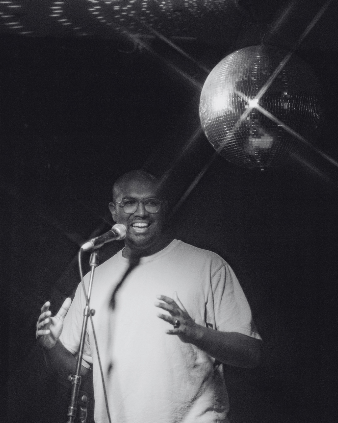 Rowan Thambar smiling and speaking into a microphone on a stage with a disco ball overhead.
